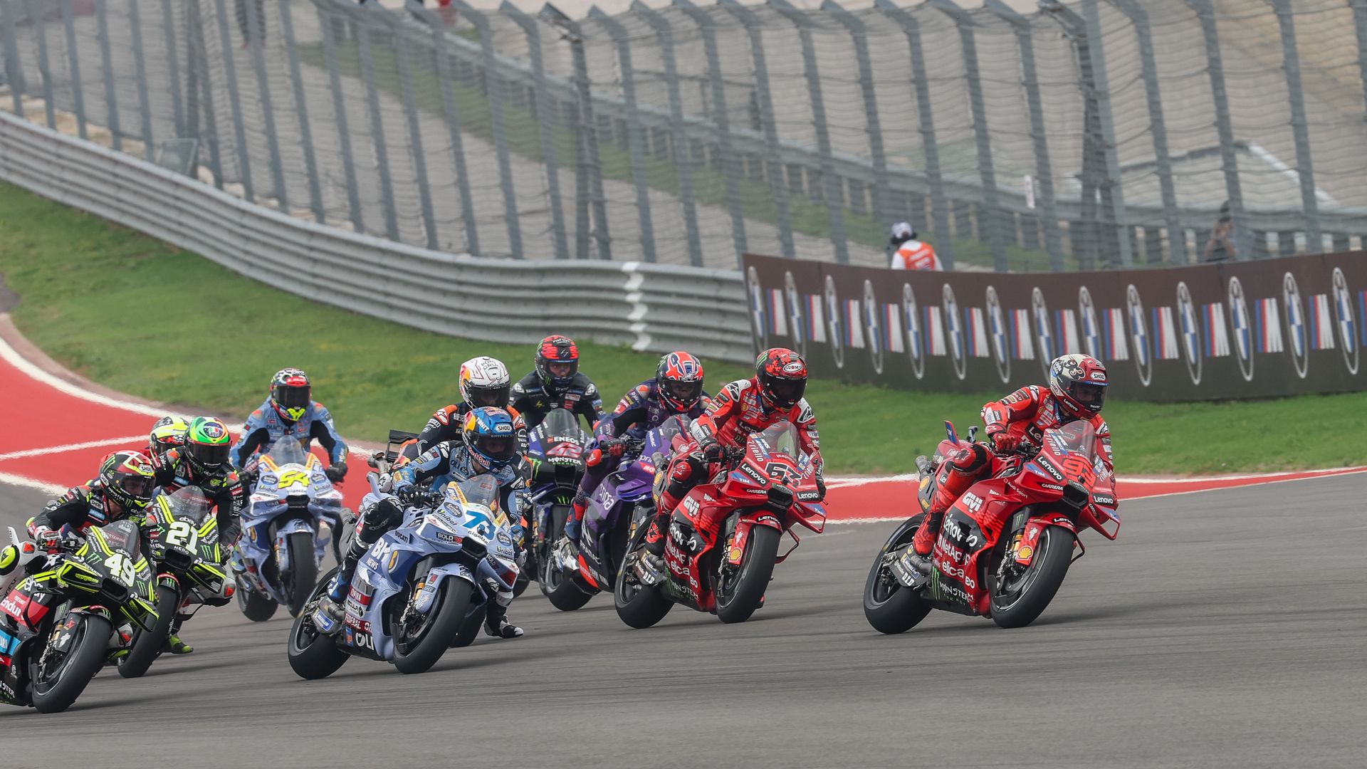 Group of motorcycle racers leaning into a curve on a race track, wearing colorful helmets and leathers. Red, blue, and black bikes sprint side by side as barriers and advertising boards line the bend.