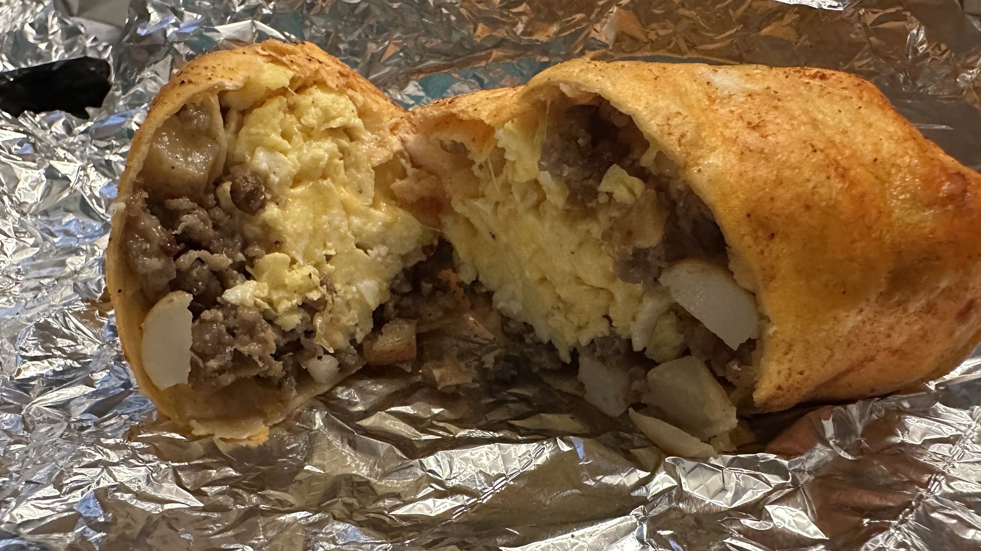 Partial burrito cut open on crinkled foil, revealing beef, mashed potatoes, and onions inside a toasted tortilla.