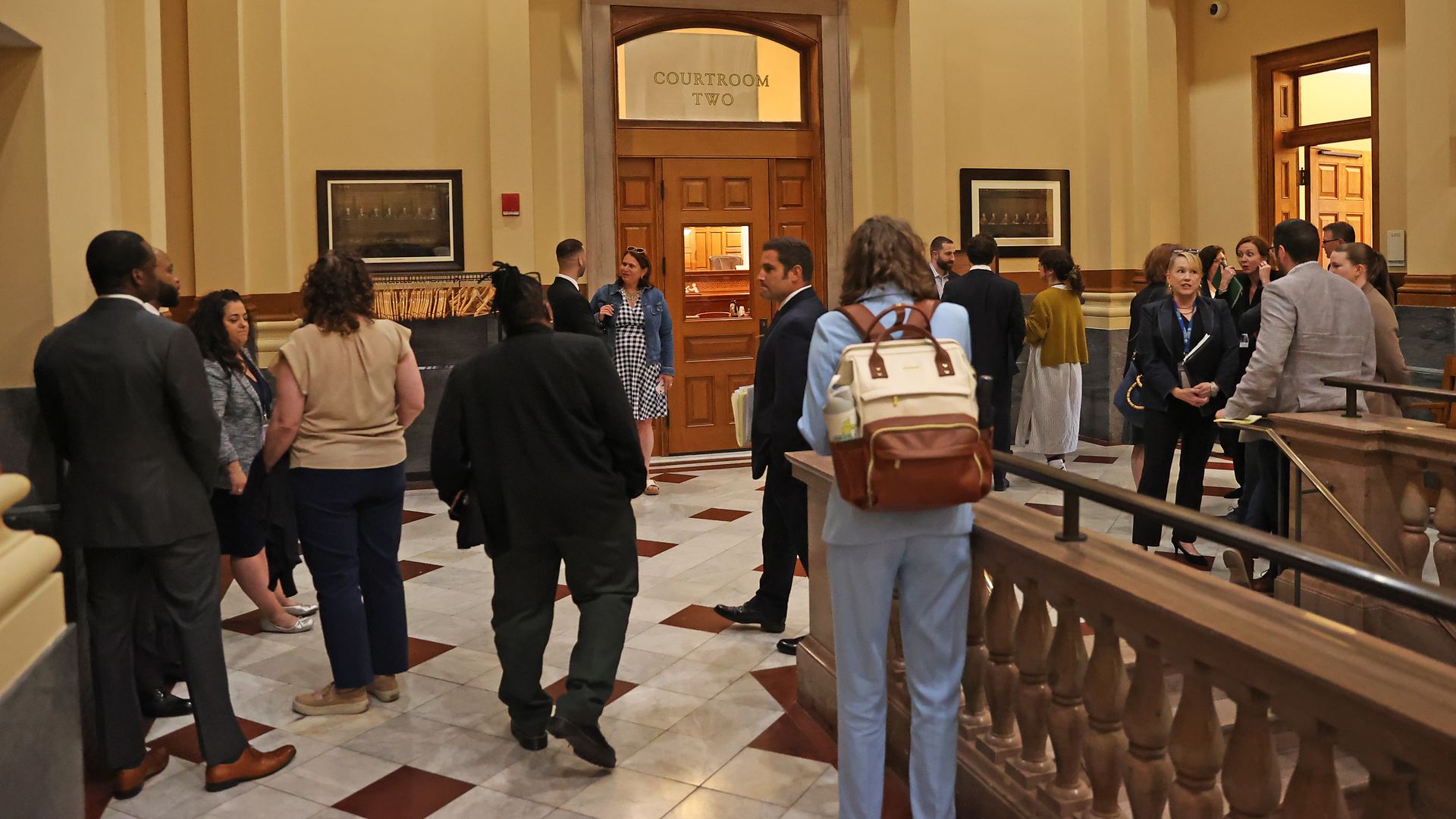People dressed in business attire gather and talk in a marble-floored hallway outside a wooden door labeled "Courtroom Two" in a building with beige walls and arches.