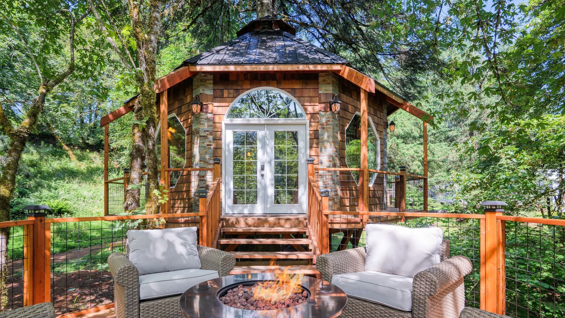Photo shows a cottage surrounded by green trees with a patio and fire pit in the foreground.