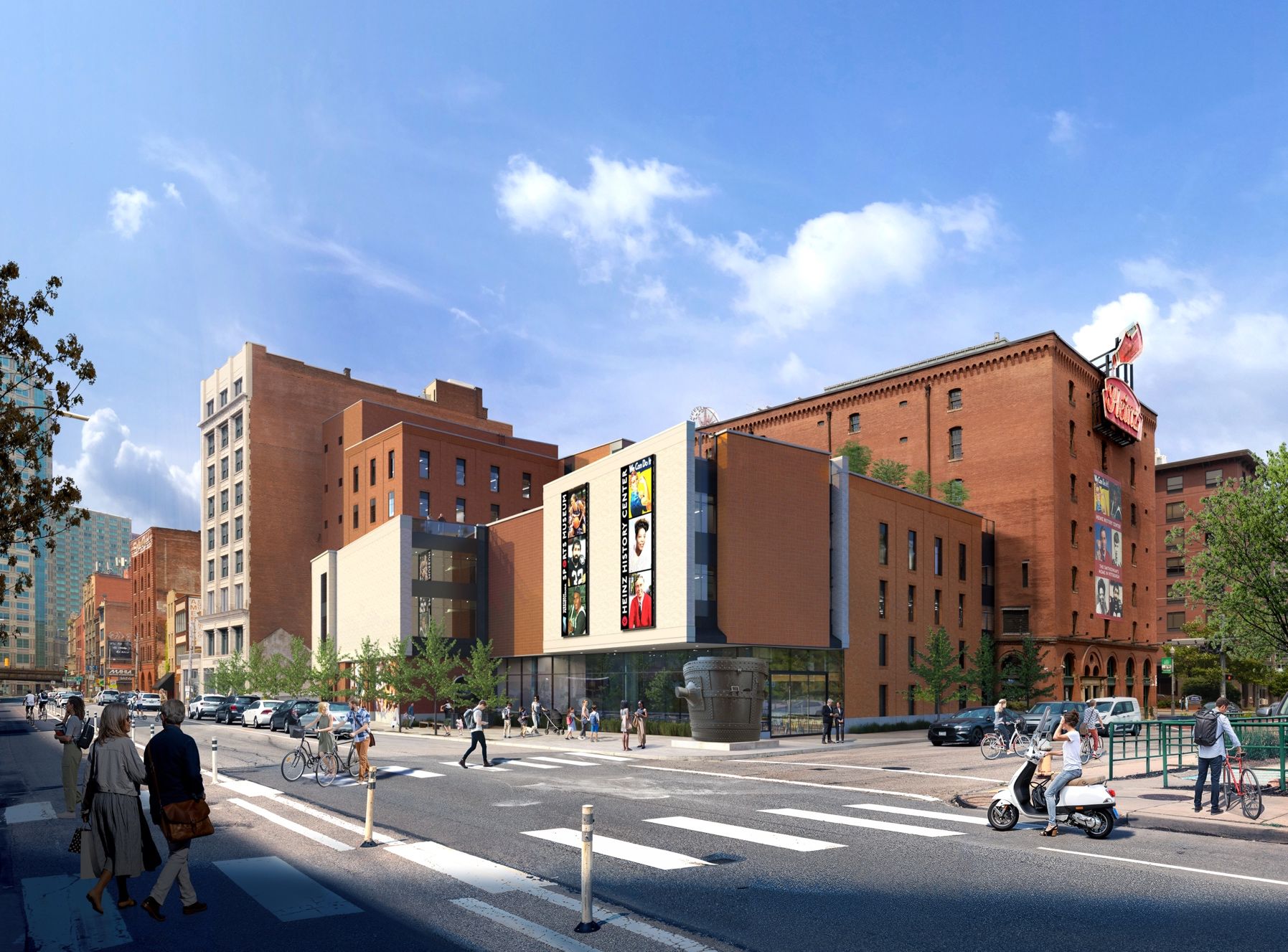 City street with red brick buildings and a modern white-beige center block featuring large poster panels. People cross the street, ride bikes, and cars drive by under a clear blue sky.