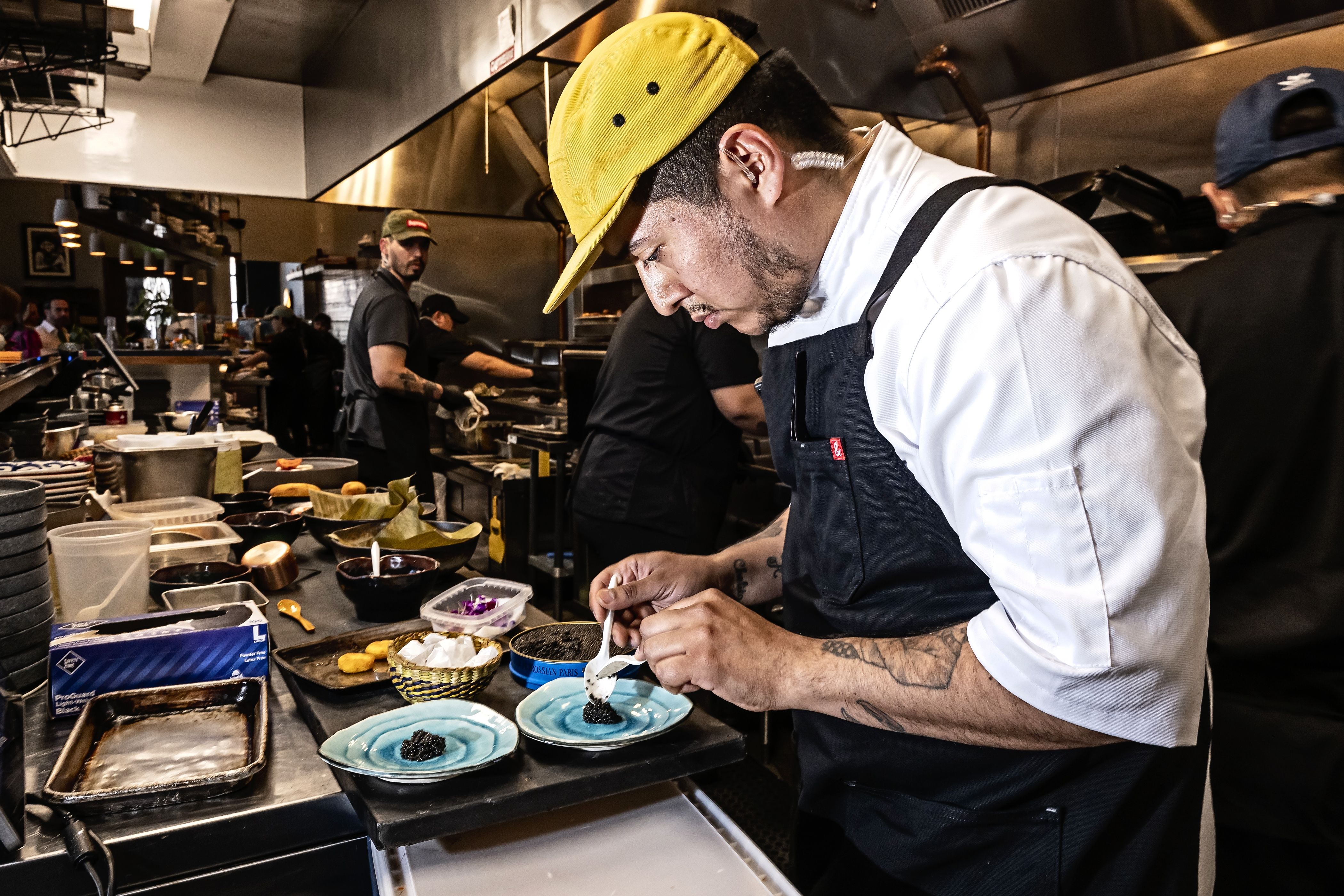 A chef in a white jacket and black apron wearing a yellow cap, intently plating purple-topped dishes in a busy stainless-steel kitchen with other cooks in the background.