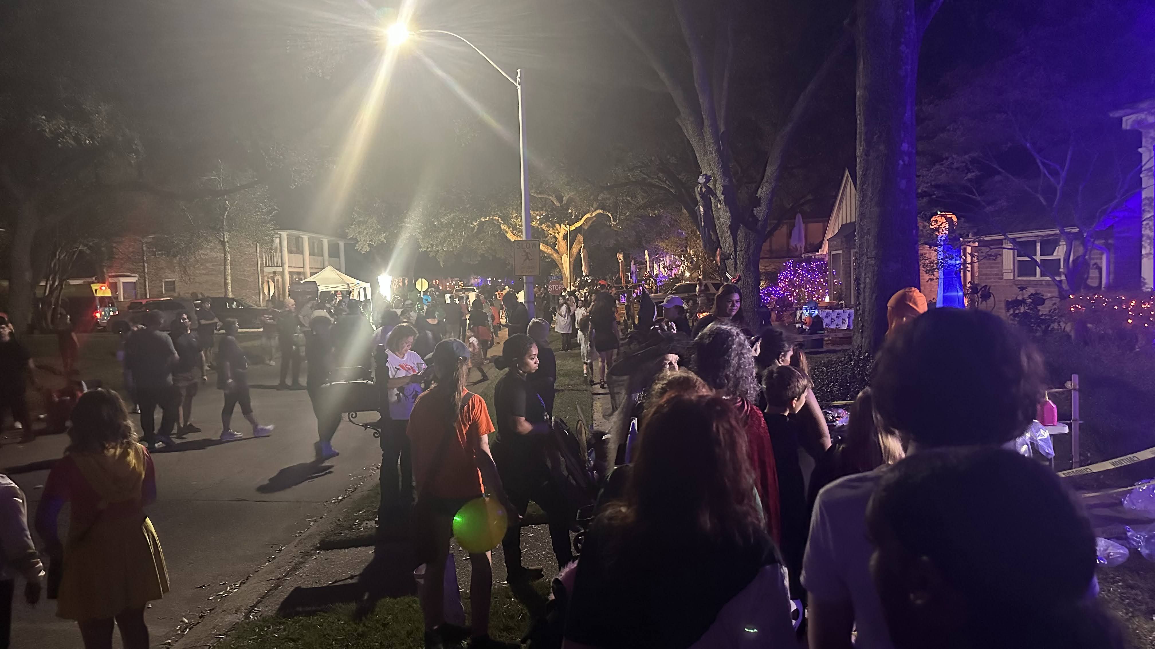 Crowd of trick-or-treaters gathered outdoors at night on a street lit by a bright streetlamp, with some colorful purple lights and home decorations visible in the background.