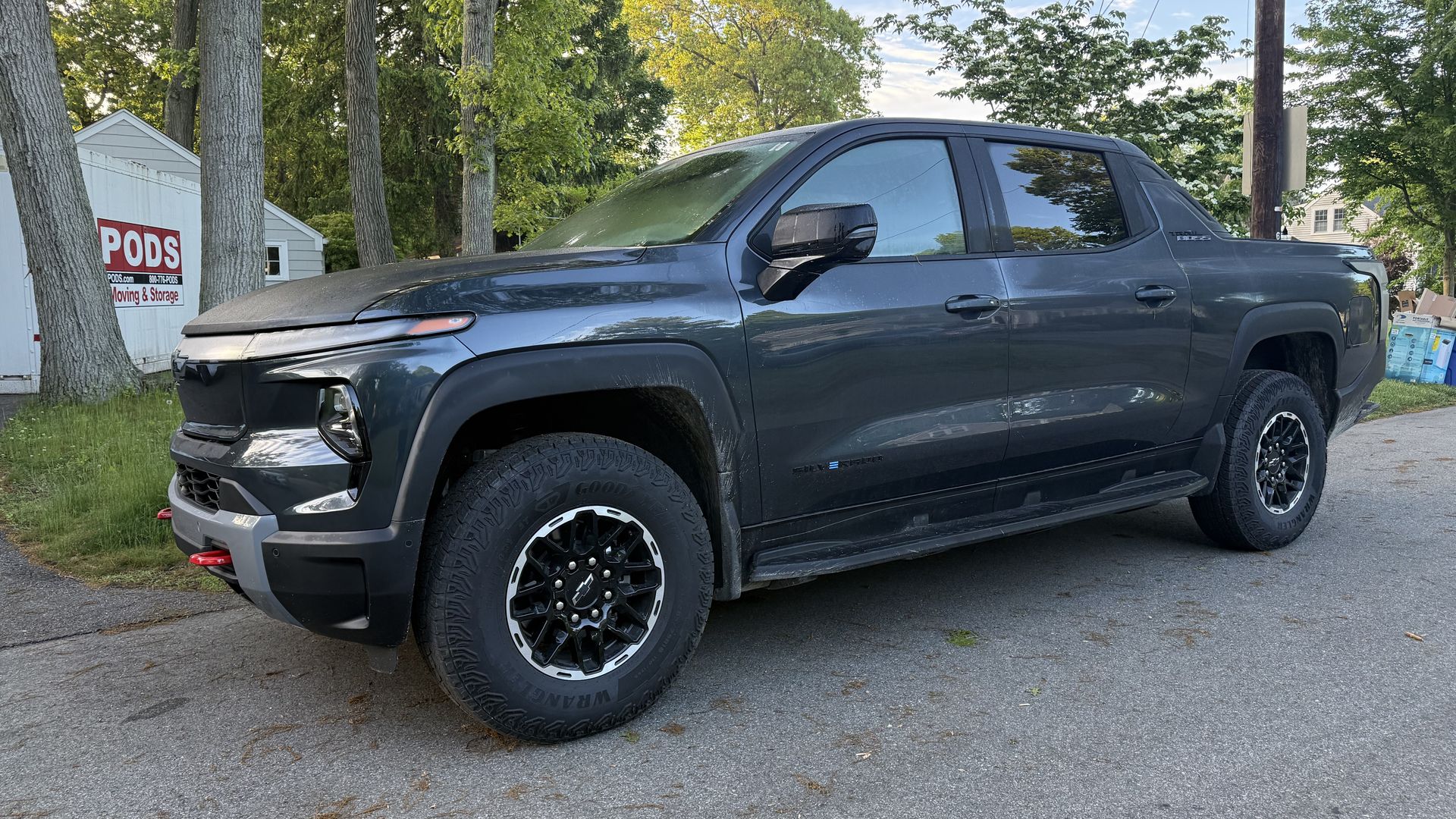 Dark gray pickup truck with black wheels parked on a suburban street. Behind it, a white PODS moving & storage shed among tall trees, with a utility pole and wires under a blue sky.