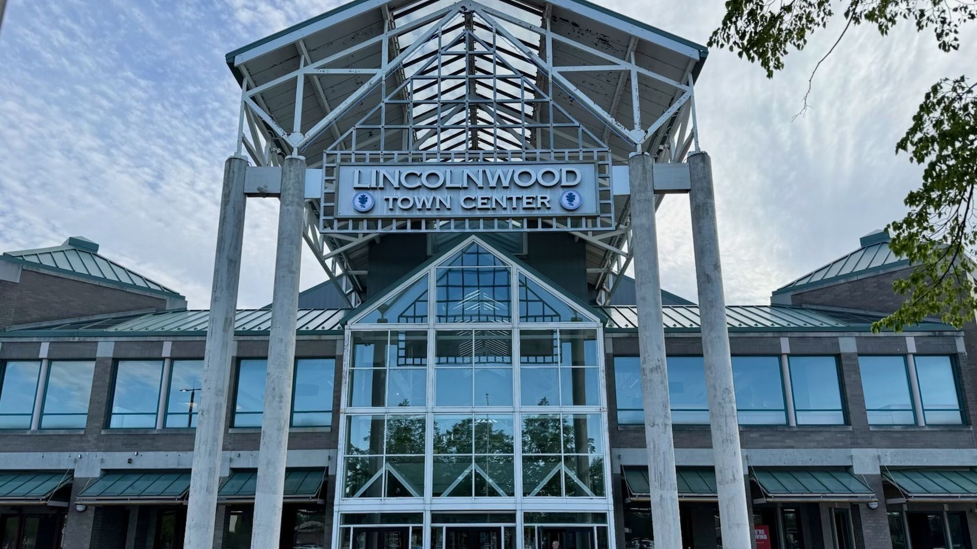Front view of Lincolnwood Town Center: glass atrium with white steel framework, tall concrete columns, brick walls, and large windows. A child in red walks on the sidewalk under a blue sky.