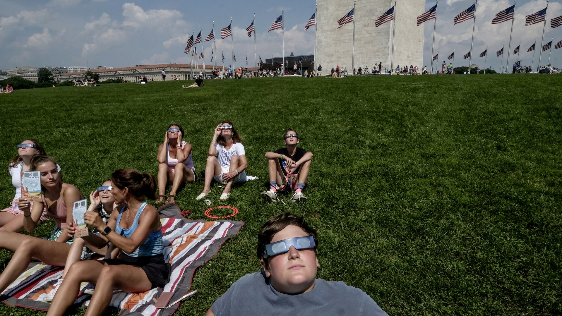 People sit in the grass in front of the Washington Monument watching an eclipse.