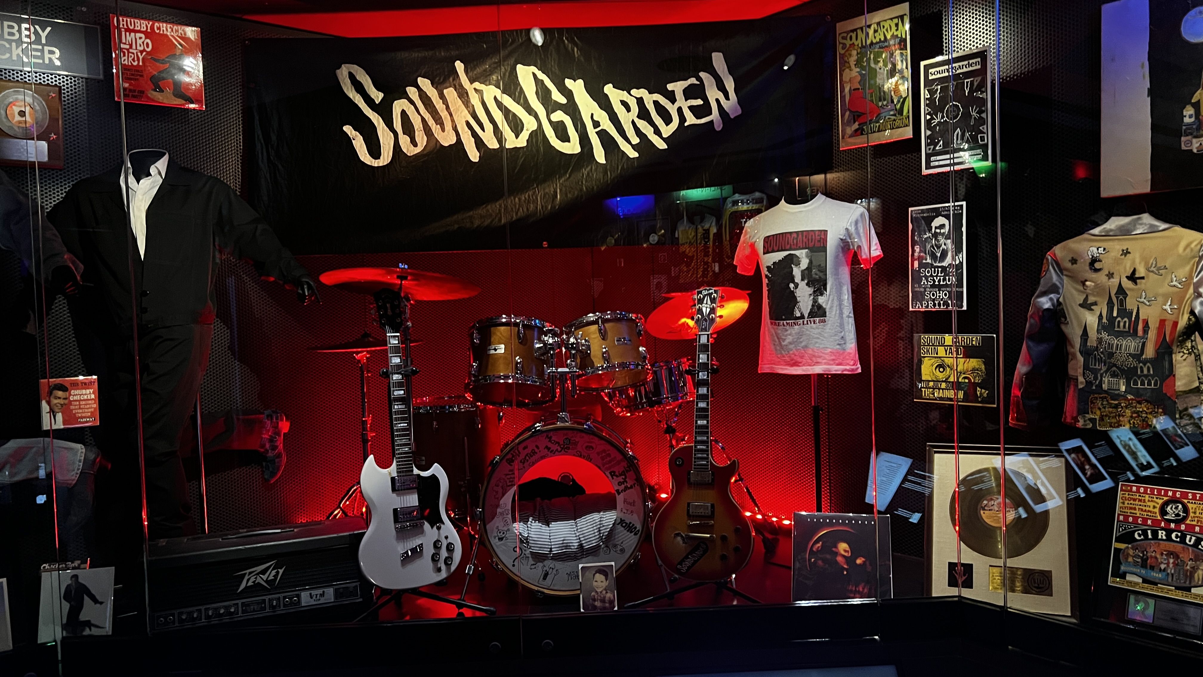 Soundgarden band exhibit featuring black banner, drum set, two guitars, white T-shirt, vintage posters, and a jacket with castle and bird designs, all showcased with red lighting.