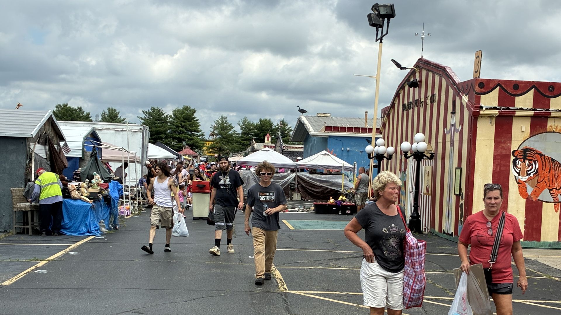 Outdoor street fair with vendor tents on the left, a striped carnival building with a tiger mural on the right, and people walking under a cloudy sky.