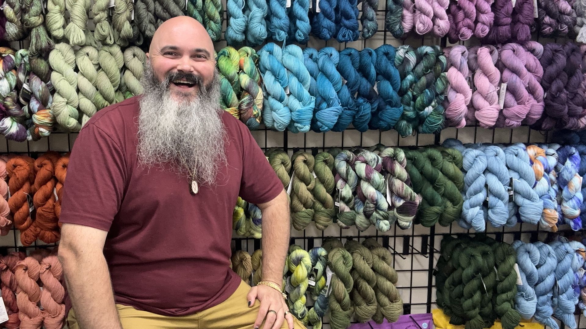 Smiling, bearded man in a maroon shirt sits in front of a wall of colorful yarn skeins arranged in rainbow gradients, with bags visible at the bottom in a yarn shop.