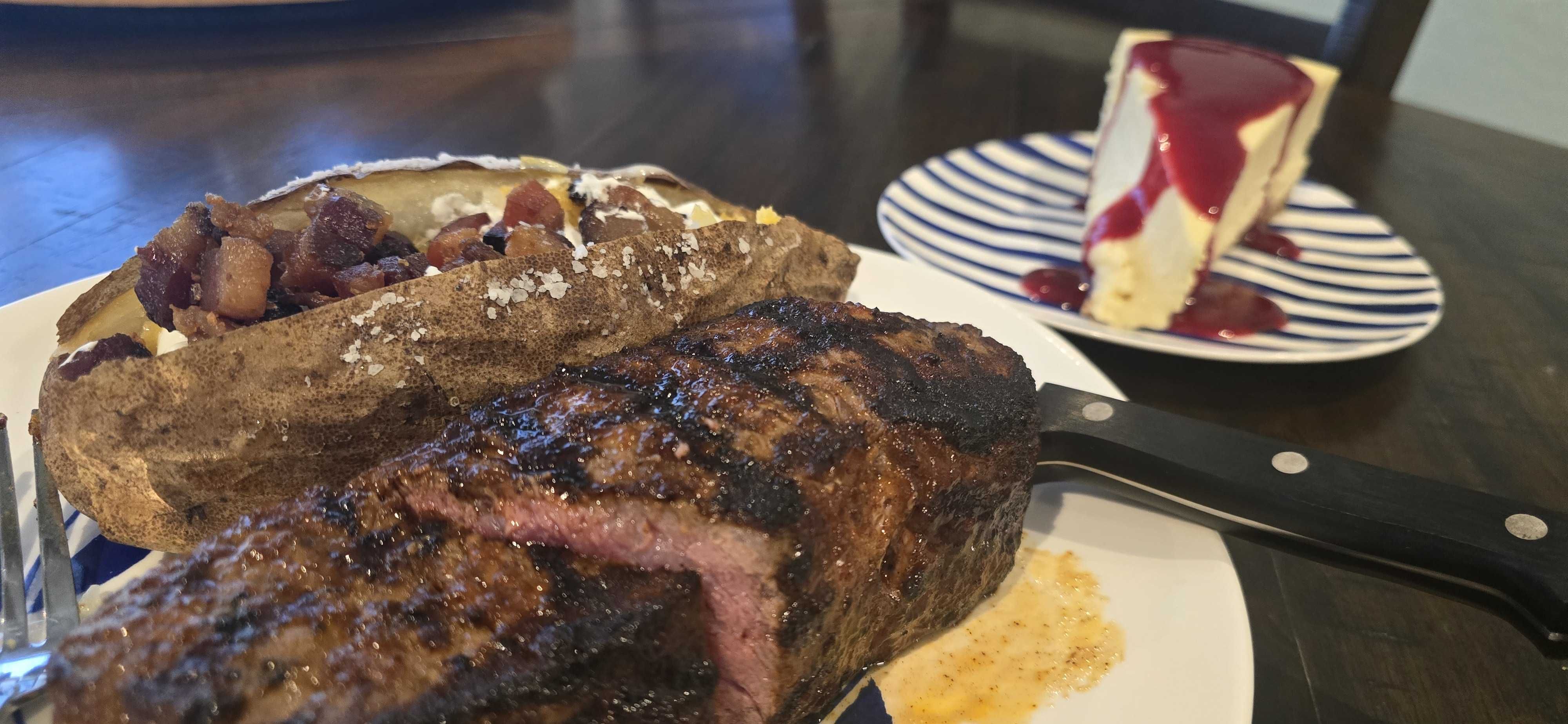 Close-up of a thick, grilled steak with a charred crust, served with a baked potato topped with bacon bits and coarse salt; a slice of berry-topped cheesecake on a striped plate is in the background.