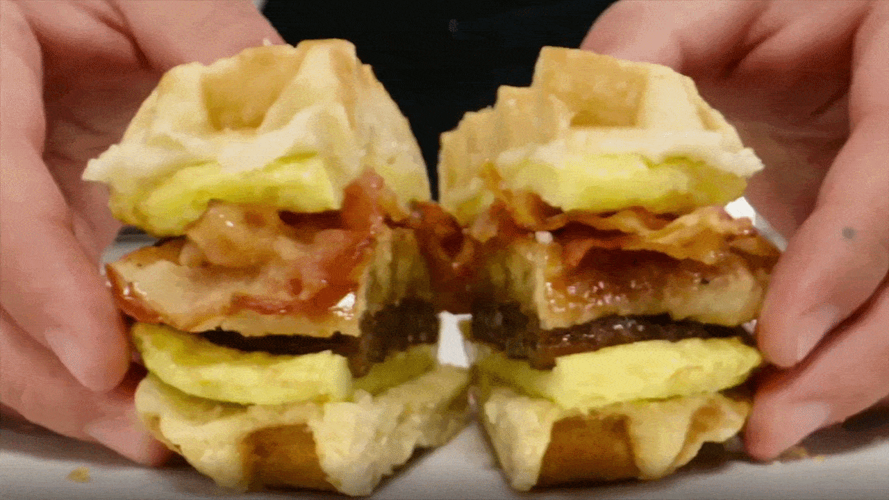 Two sandwich halves held by hands are being pulled apart, revealing bacon, a meat patty, and pale yellow fillings between light buns.