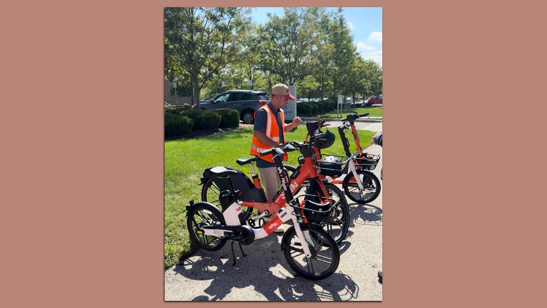 A man wearing an orange safety vest and a pink cap stands beside several red Spin rental bicycles with front baskets on a sunny sidewalk, with green grass, trees, and parked cars in the background.