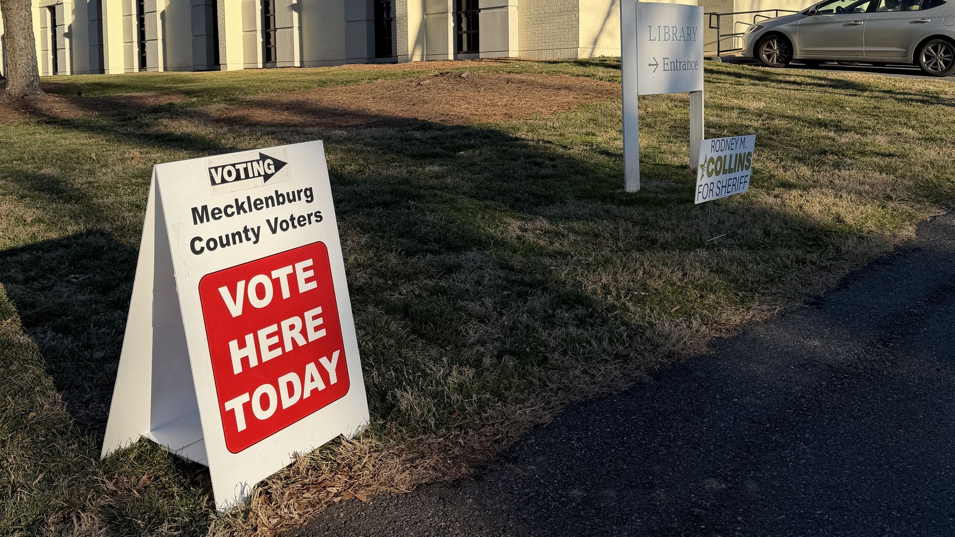 Beige library exterior with a silver car parked on the right; foreground shows election signs reading "VOTE HERE TODAY" and "Mecklenburg County Voters" and a "LIBRARY Entrance" sign.