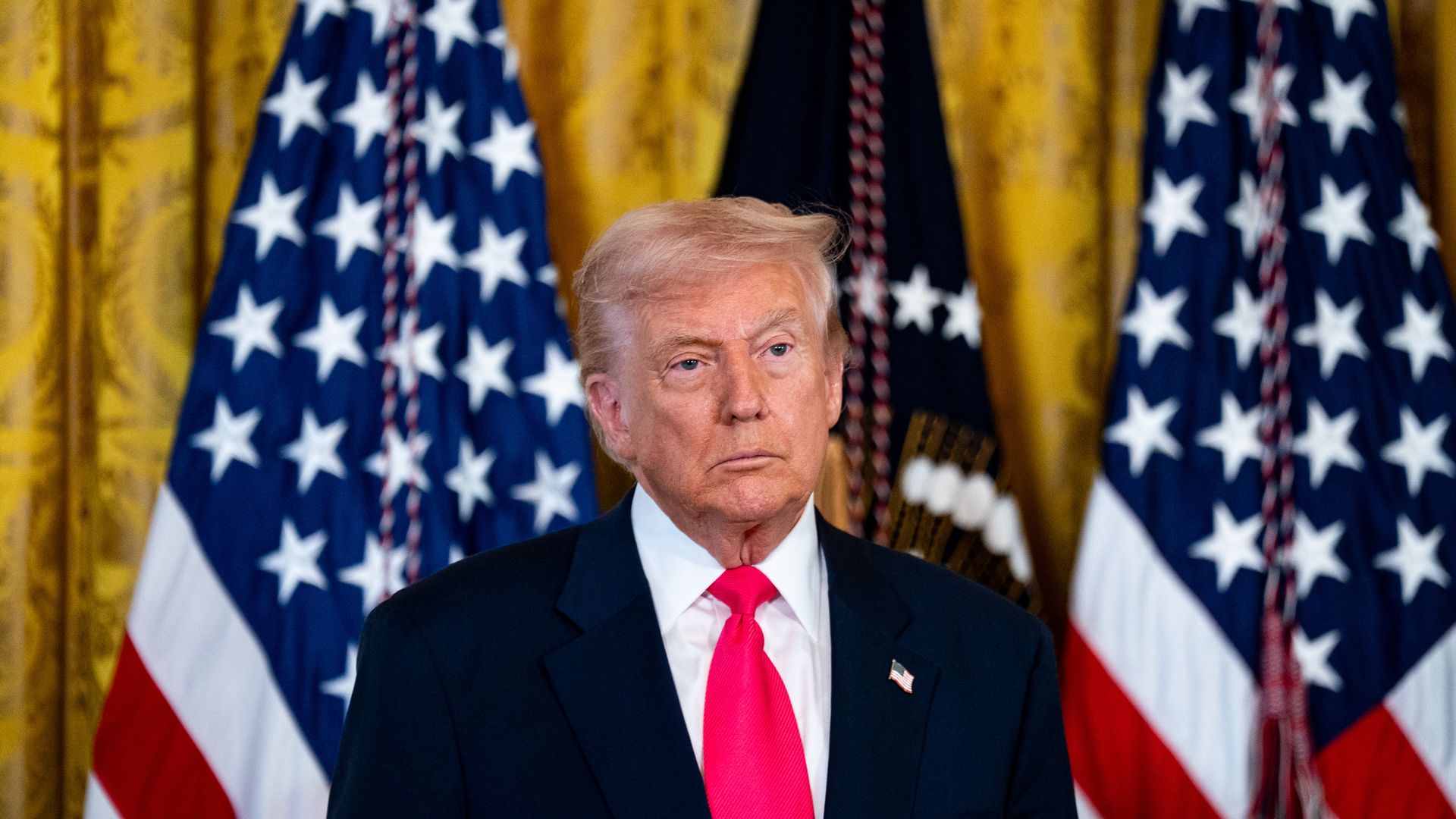 Trump, wearing a dark suit, white shirt and red/pink tie, stands in front of American flags 