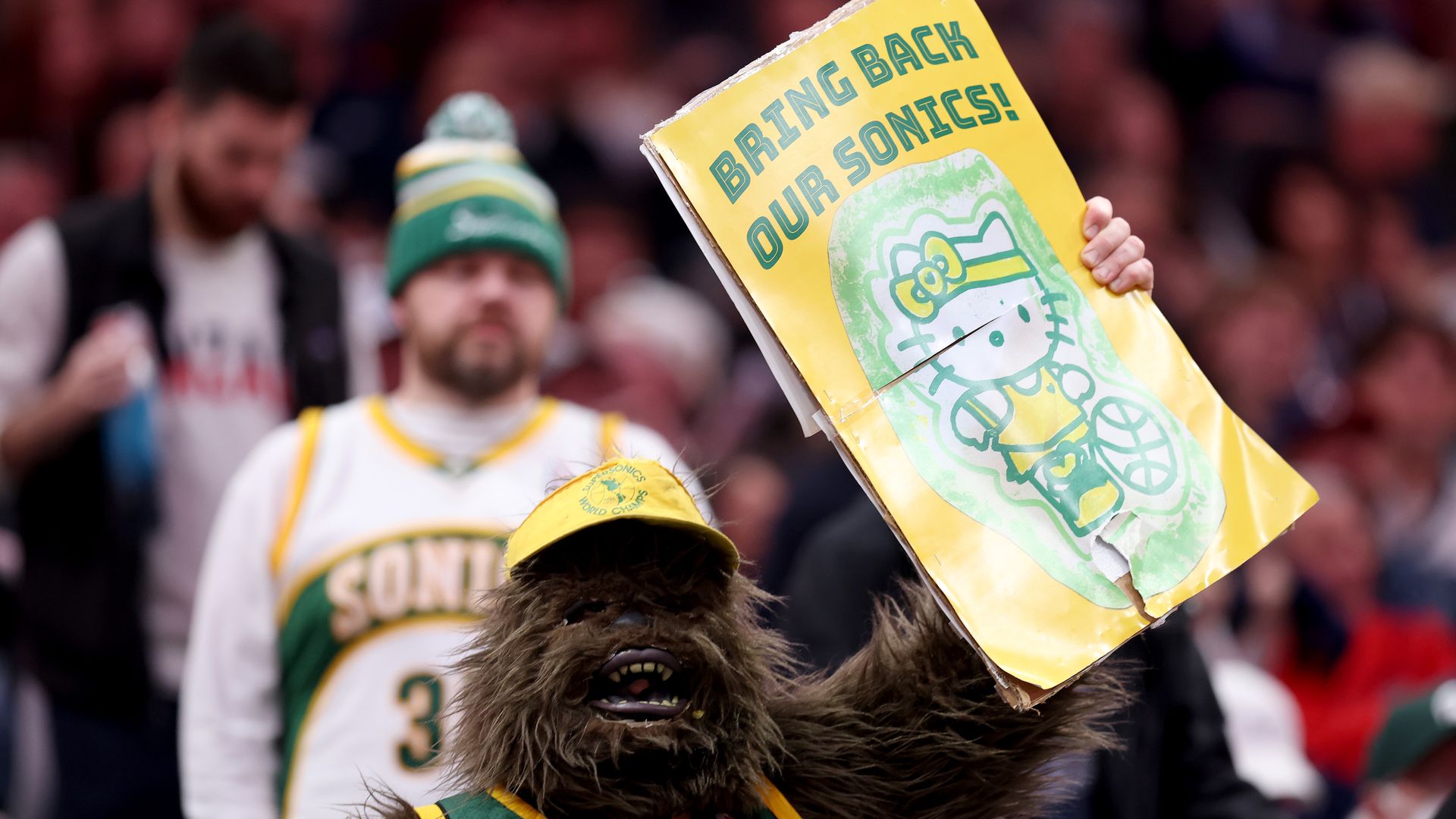 A person in a sasquatch costume holds up a sign that says "Bring back our Sonics."