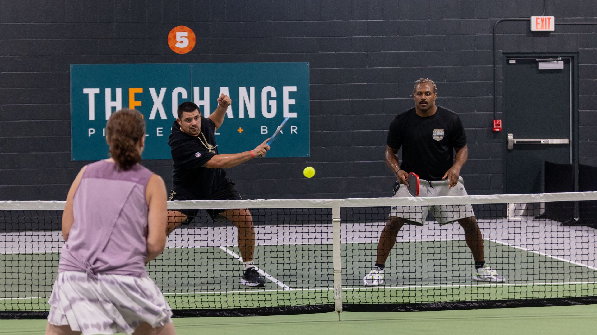 Three adults playing pickleball indoors on a green court with a net and a gray back wall featuring a sign reading "THE EXCHANGE." Two men face one woman, hitting a yellow ball.