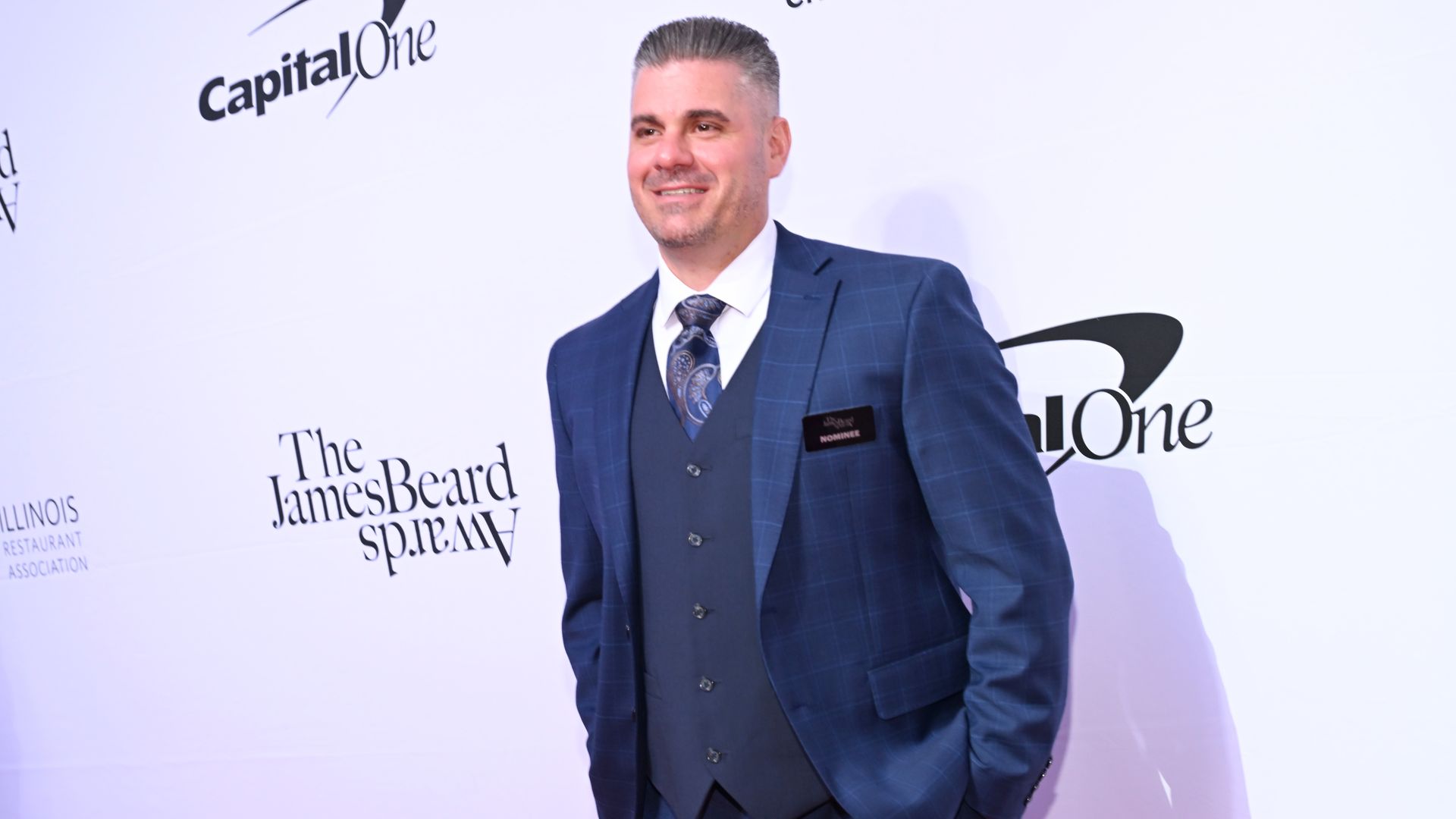 Man in a blue plaid suit, white shirt, and paisley tie posing at The James Beard Awards event with Capital One logo on backdrop, wearing a nominee name tag.
