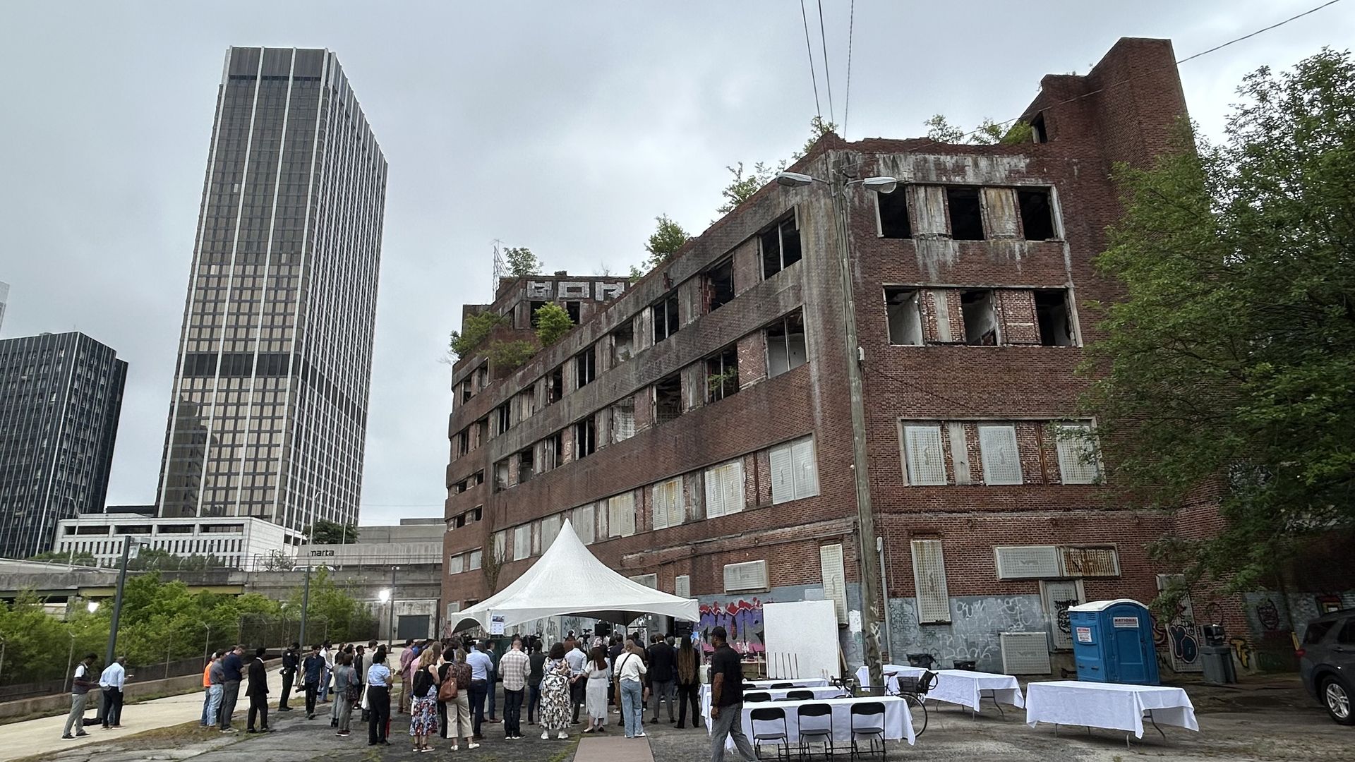 A large group of people under and near a large canopy tent stand outside a dilapidated building with trees growing from the windows and roof