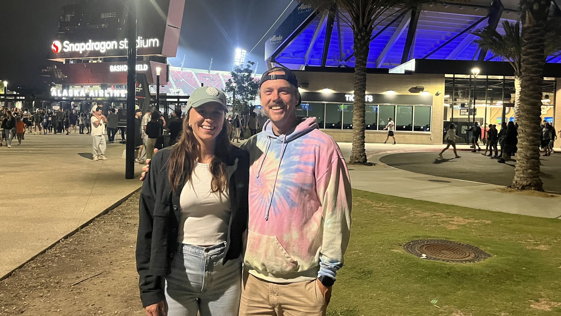 Smiling man in tie-dye hoodie and woman in baseball cap and casual clothes posing at night outside Snapdragon Stadium with crowds, palm trees, and bright lights in the background.