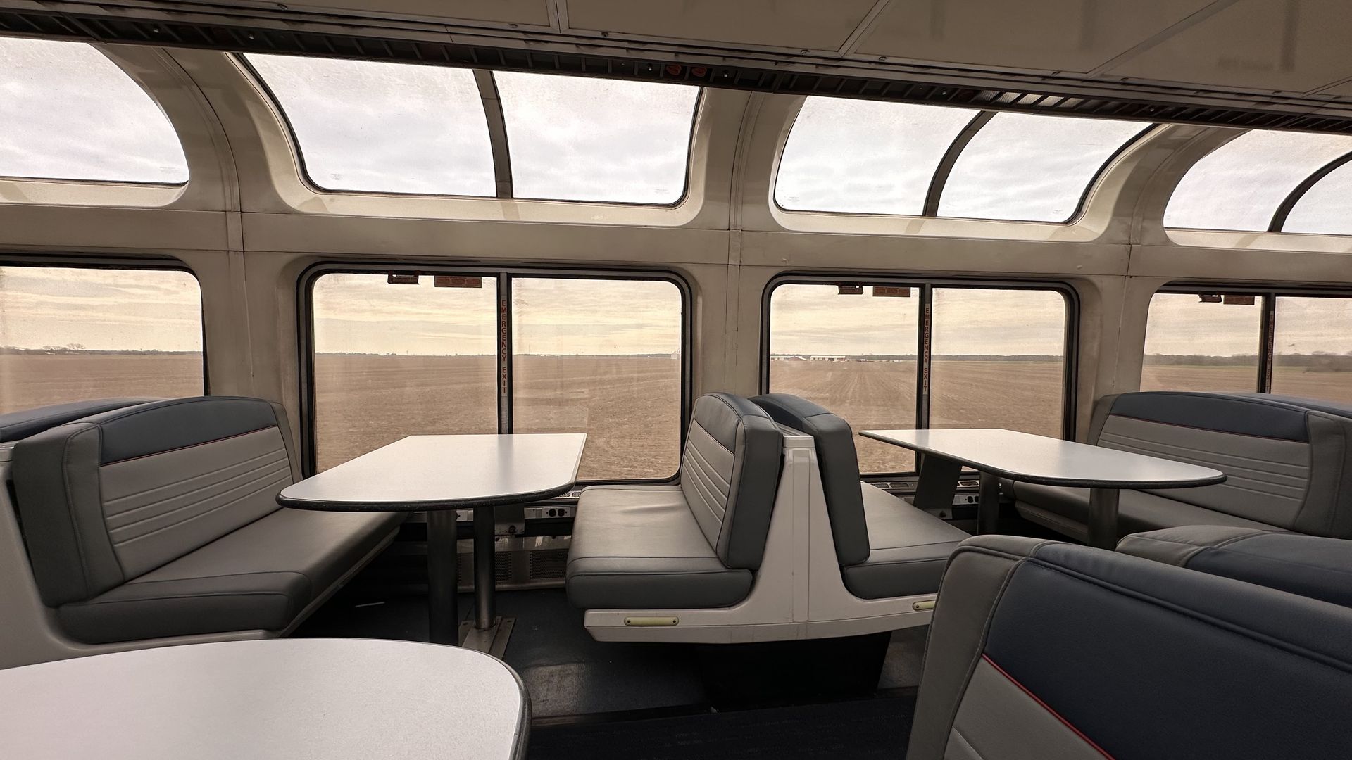 Interior of the Texas Eagle Amtrak en route to Chicago from Dallas. There are gray booth seats and white tabletops; large curved windows reveal flat farmland under an overcast sky.