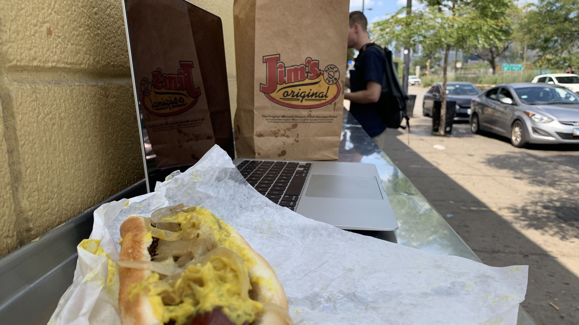 Outdoor street scene with a laptop on a reflective metal table, a bag labeled "Jim's Original", and a hot dog with mustard. A person with a backpack stands nearby as cars and trees line the street.