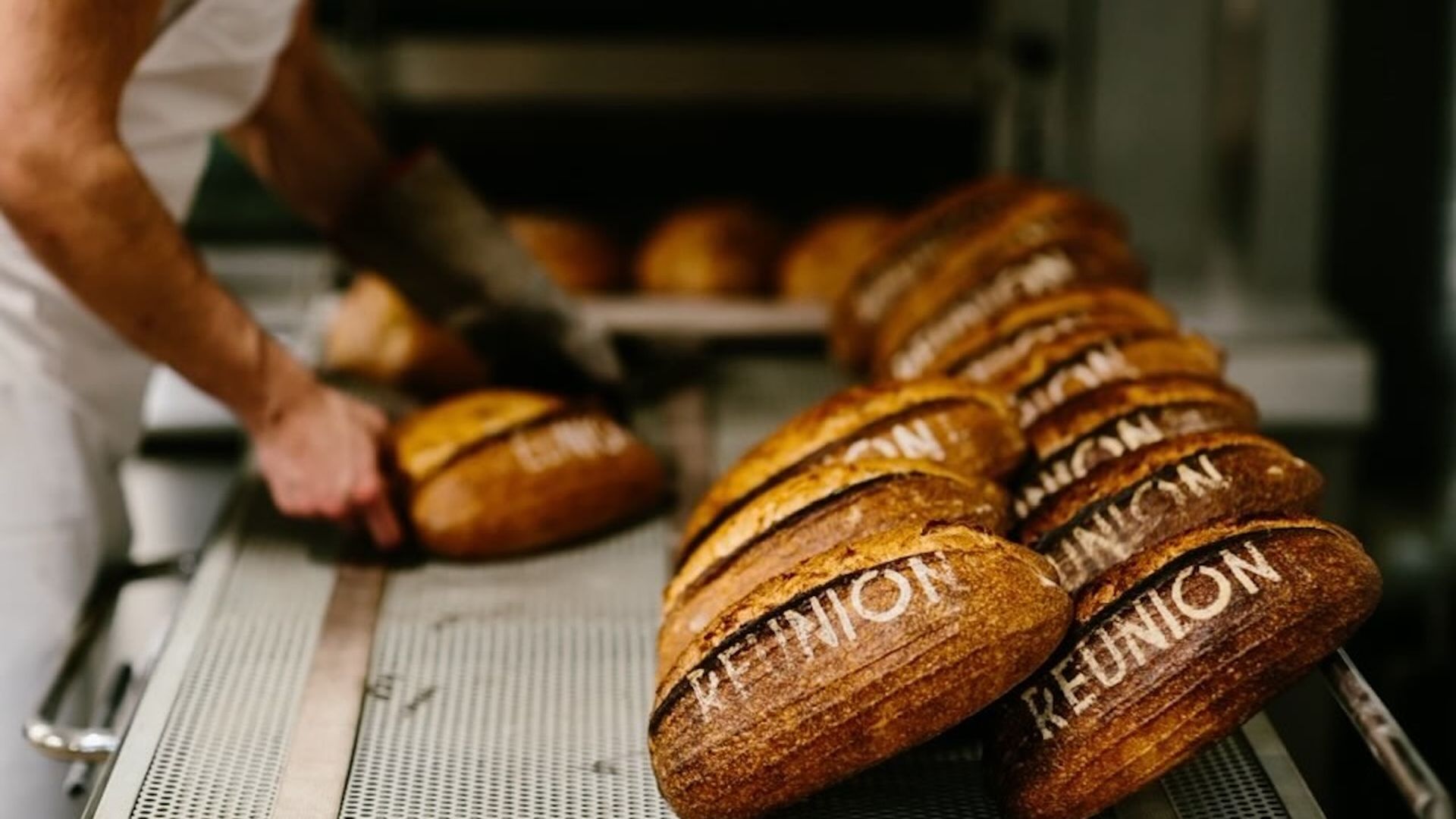 Row of round brown loaves stamped with "UNION" on a bakery conveyor belt, while a baker slices another loaf in the background.