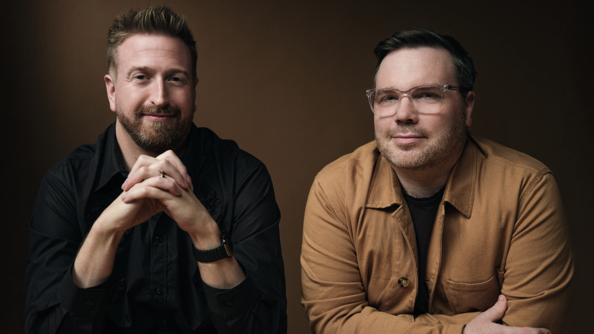 Two men sit at a brown studio backdrop, smiling at the camera. Left man in a black shirt with clasped hands; right man wears a tan jacket and clear glasses.
