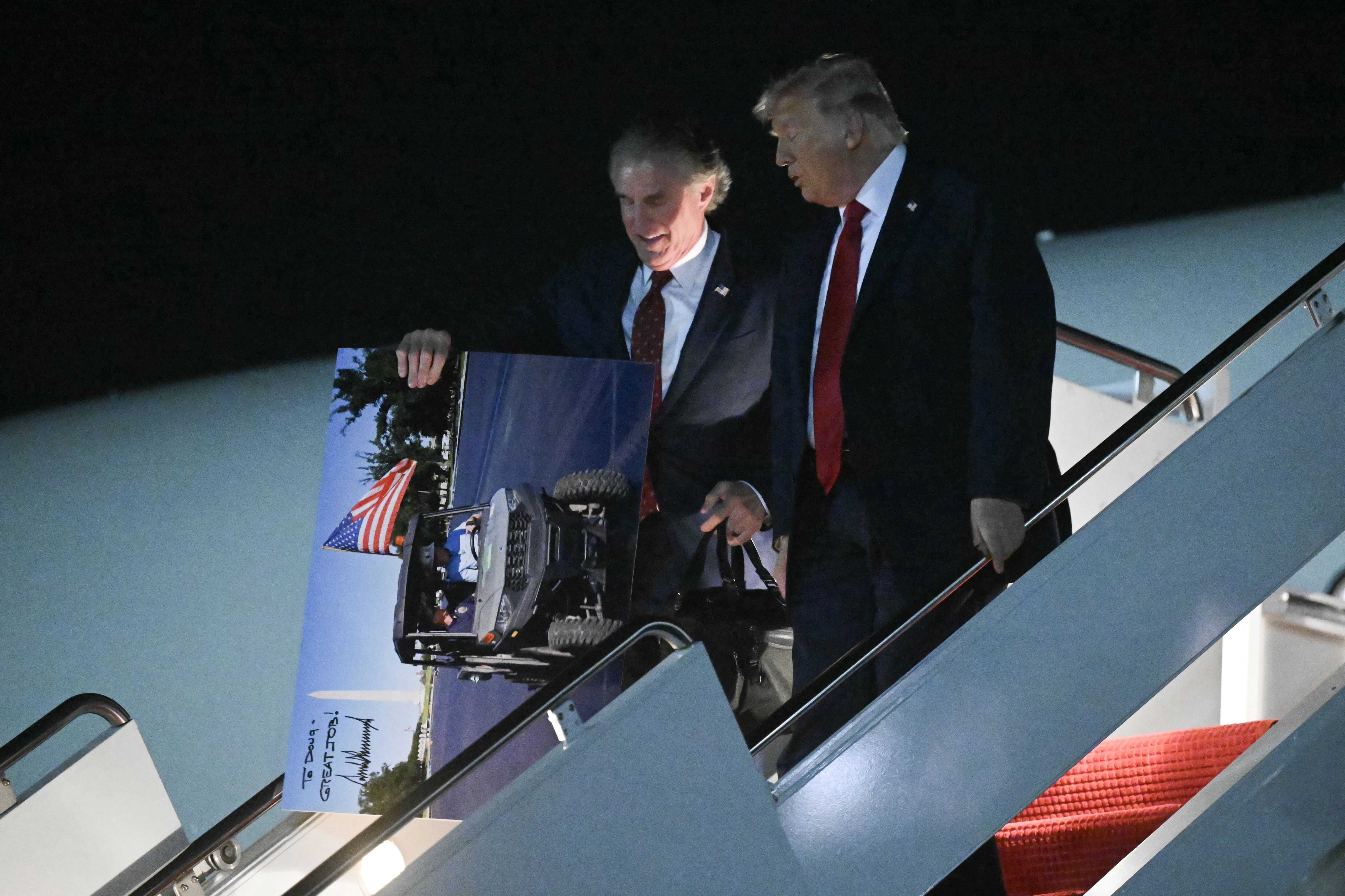 While disembarking from Air Force One next to President Trump, Doug Burgum holds a giant poster of a golf cart riding on a freshly painted surface of the Reflecting Pool