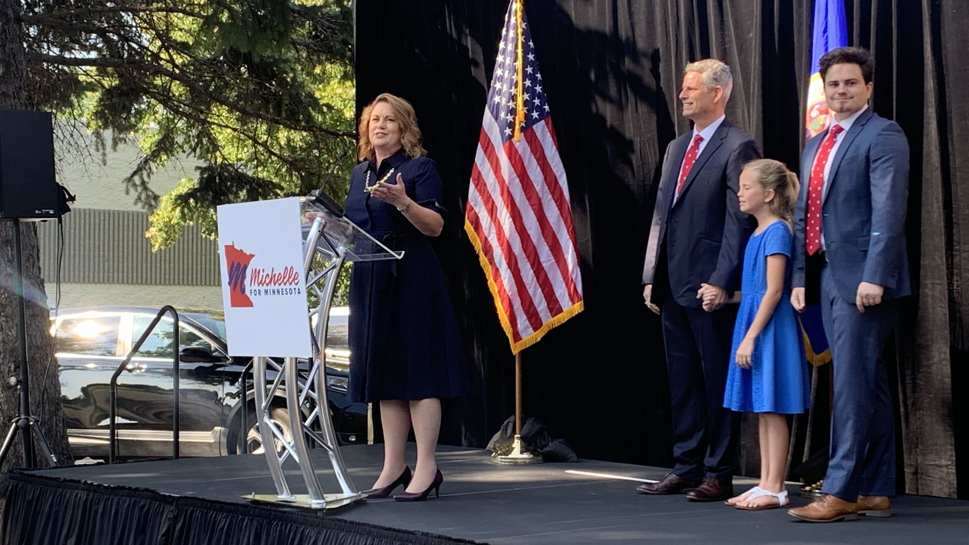 Michelle Benson speaks at a podium alongside her husband and two children.