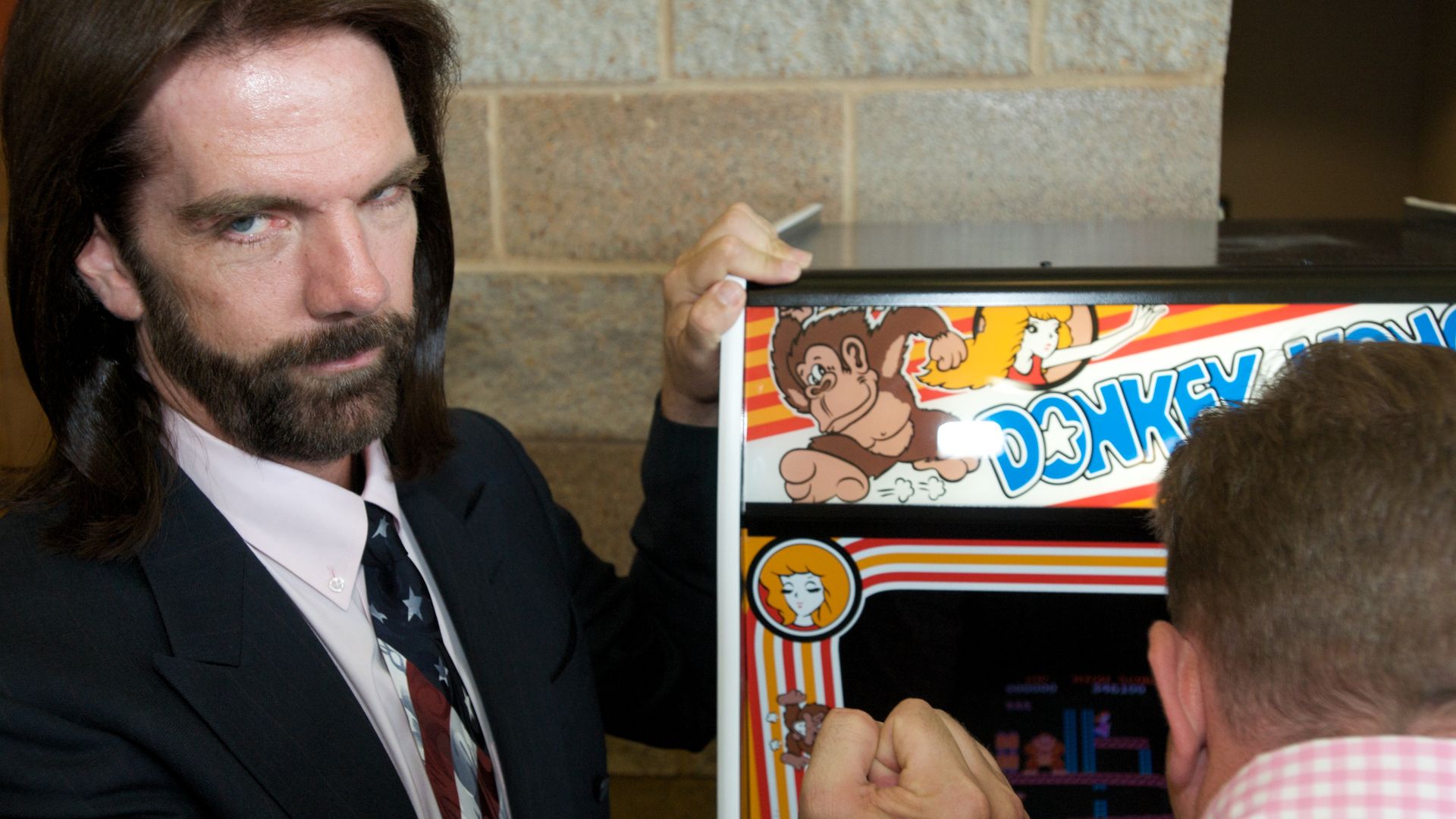 Photo of a man in a suit making a fist while he stands next to a Donkey Kong machine