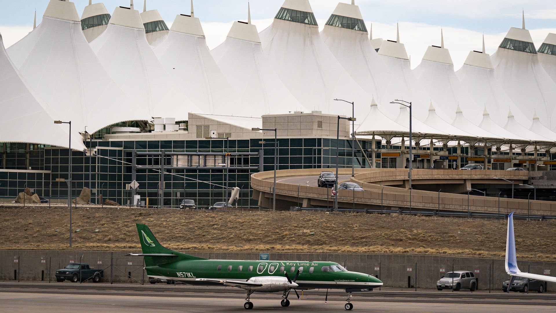 An airport terminal with white, tent-like roofs and glass walls; a green Key Lime Air regional aircraft sits on the tarmac in the foreground, with fencing, vehicles, and a curved roadway nearby.