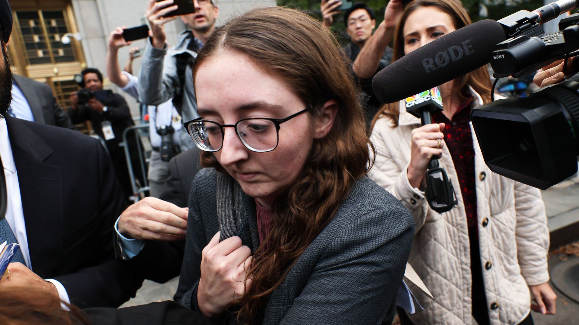 A woman at the center of the frame is huddled down as reporters and cameras look on. She is wearing a grey coat with long brown hair and glasses. 