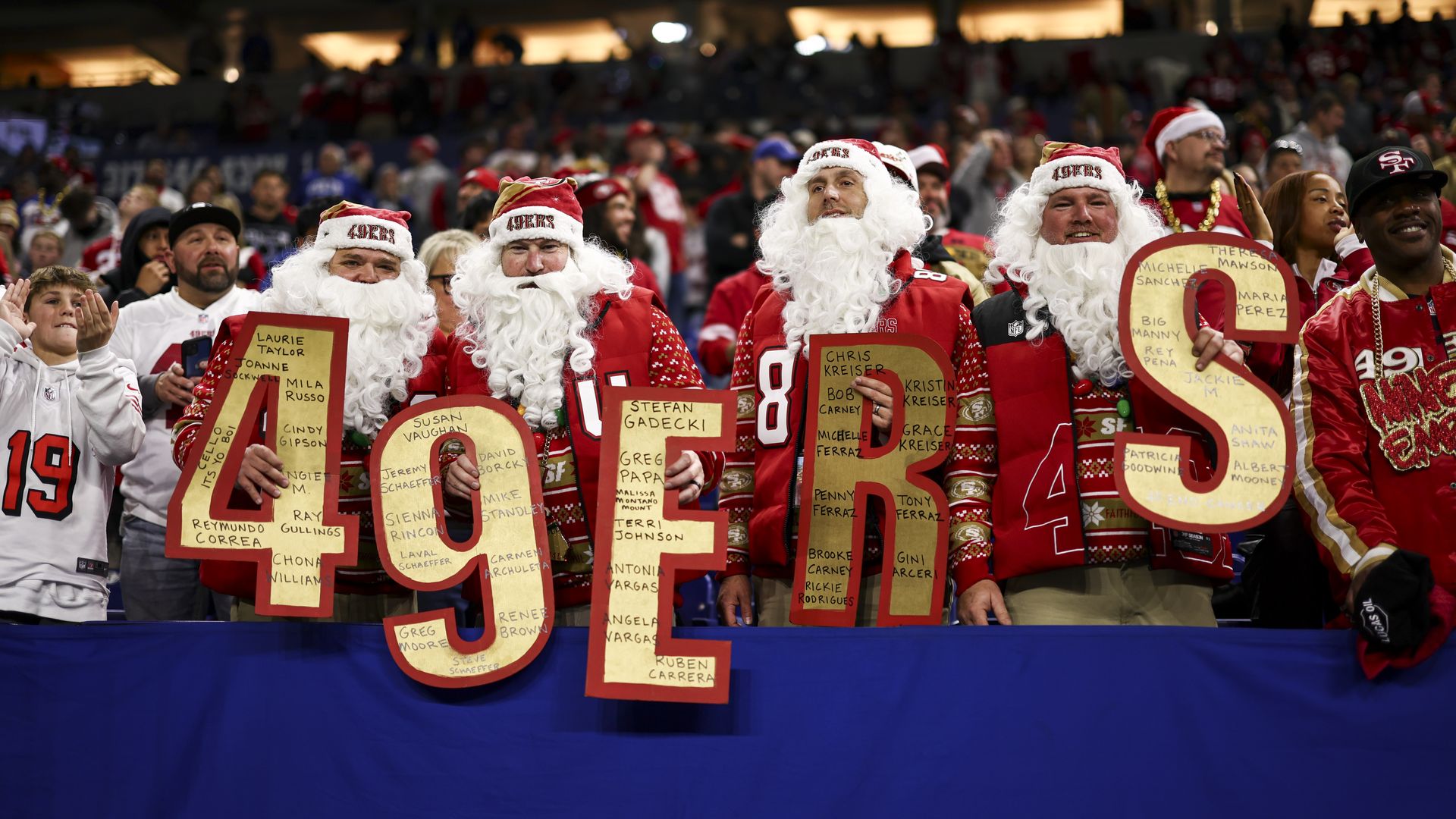 Four men dressed as Santa Claus with white beards and red 49ers hats hold large red and gold letters spelling "49ERS" at a crowded football stadium.