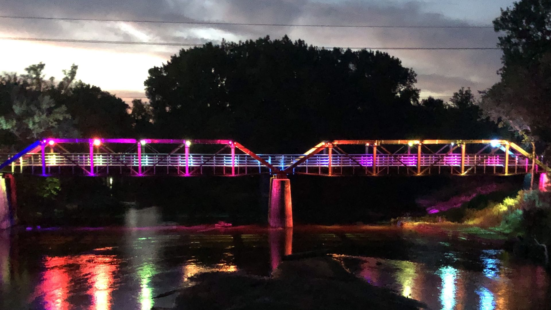 Evening scene of a neon-lit pedestrian bridge over water, reflections of pink, purple, and blue lights shimmering on the surface. Dark silhouetted trees and a cloudy sky with power lines overhead.