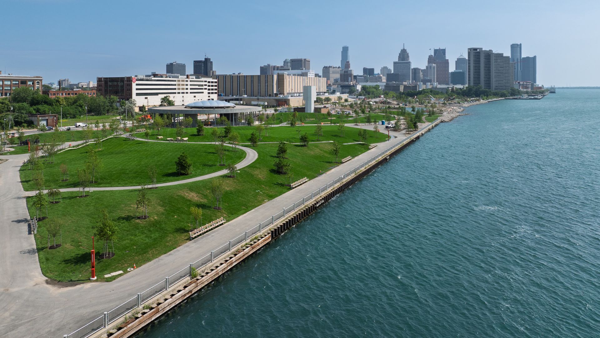 Waterfront park with green lawns, paths, and benches along a blue lake, city skyline with high-rise buildings in the distance, under a clear blue sky.