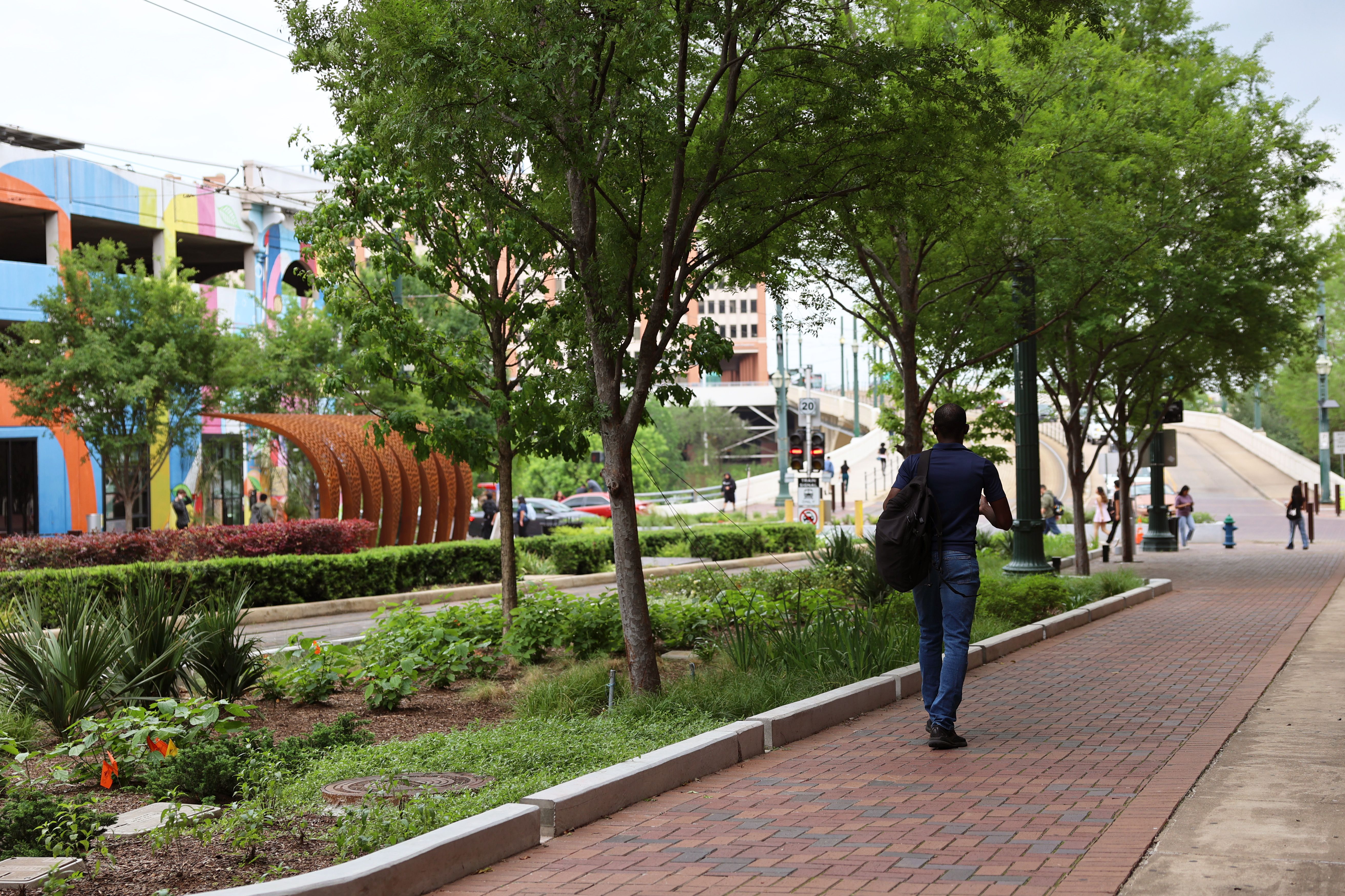 Person with a backpack walks along a brick sidewalk through a tree-filled city park; colorful mural walls on the left and an orange curved sculpture in the foreground.