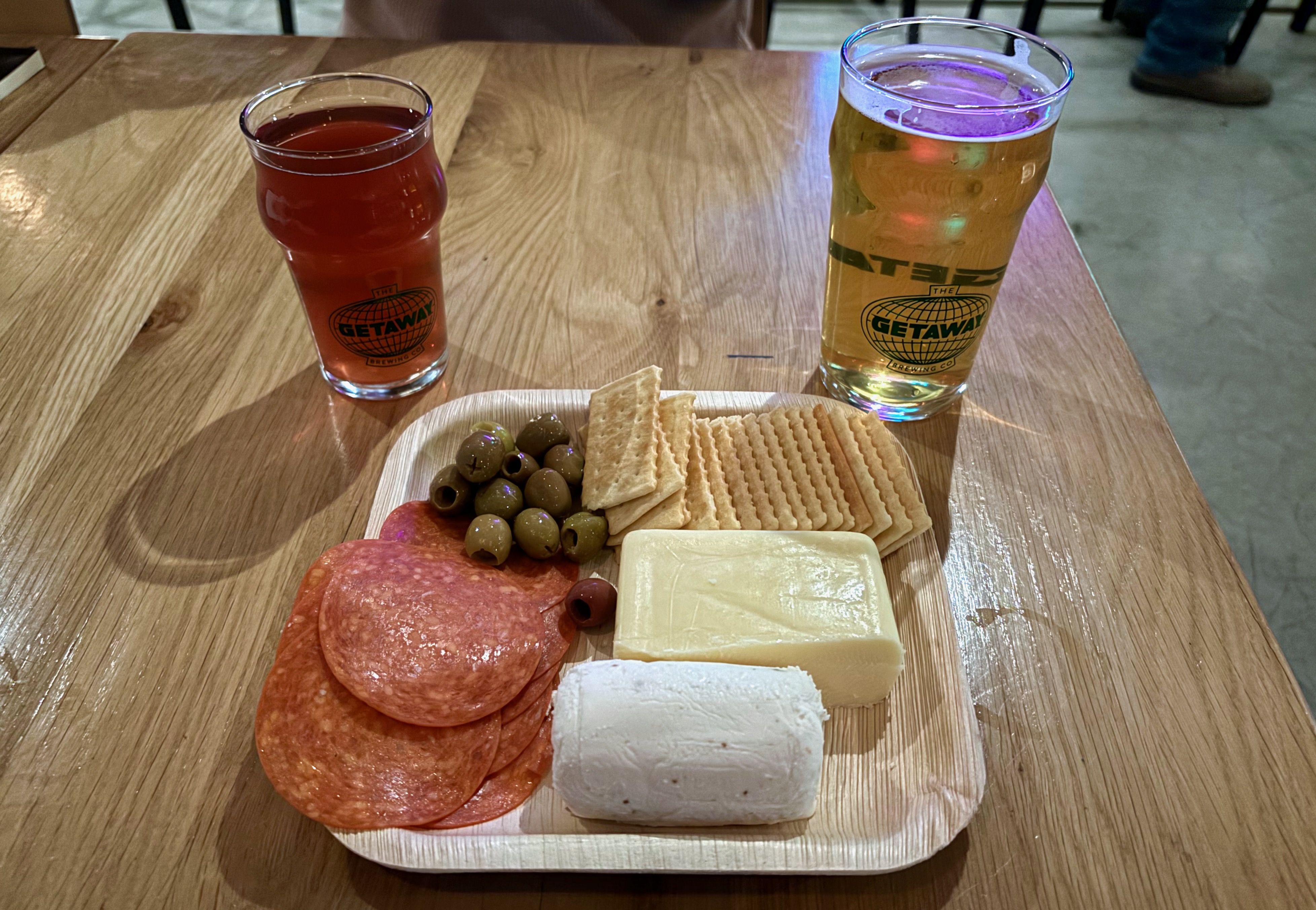 A charcuterie board featuring pepperoni, olives, crackers and two types of cheeses next to two beer glasses