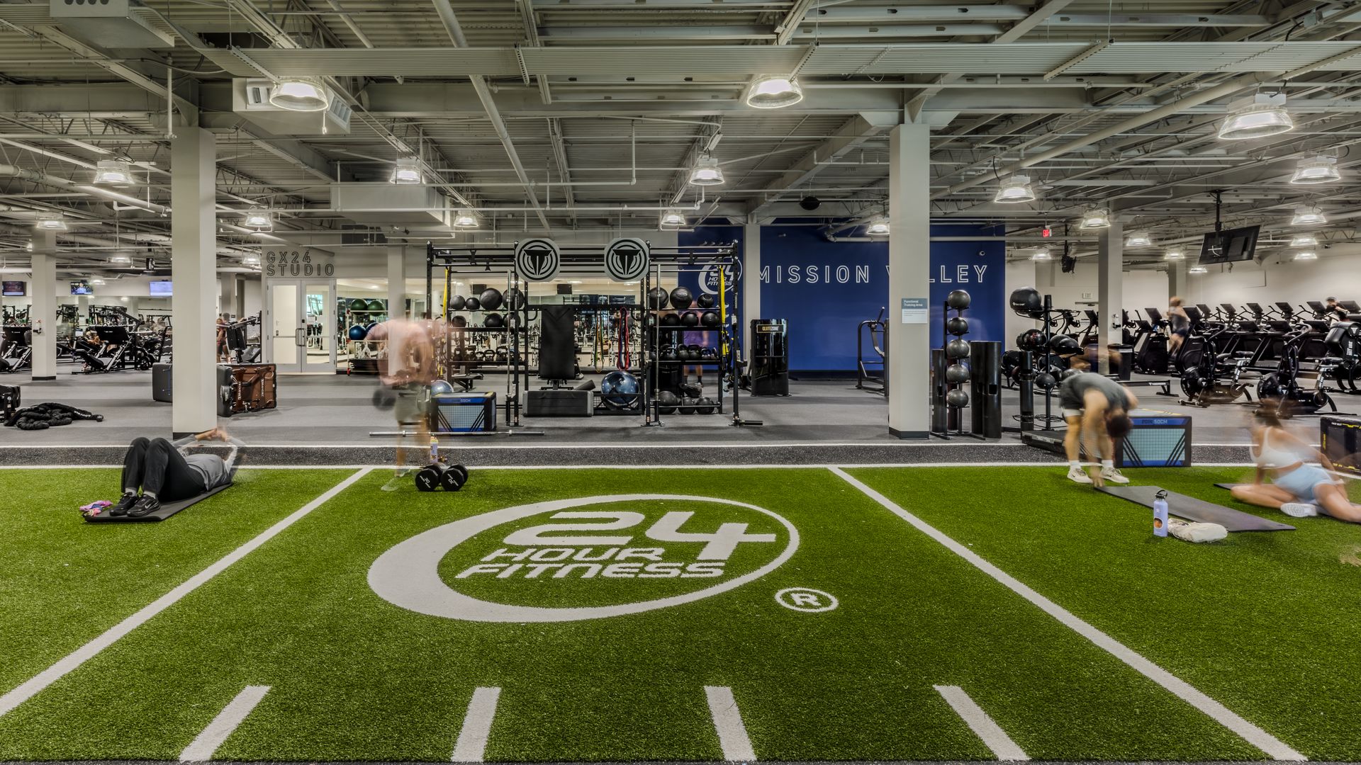 Indoor gym with a large turf area in the foreground marked by a white circle; people exercise on mats and weights, with cardio machines along the back and a blue wall reading "MISSION VALLEY".