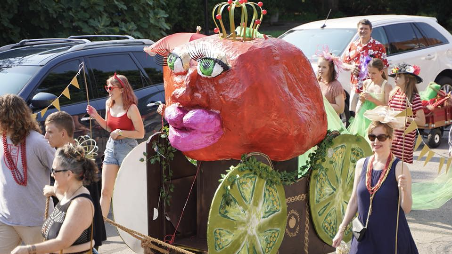 A colorful parade scene featuring a giant red tomato sculpture with a gold crown on a wheeled float, surrounded by people in bright outfits and festive accessories.