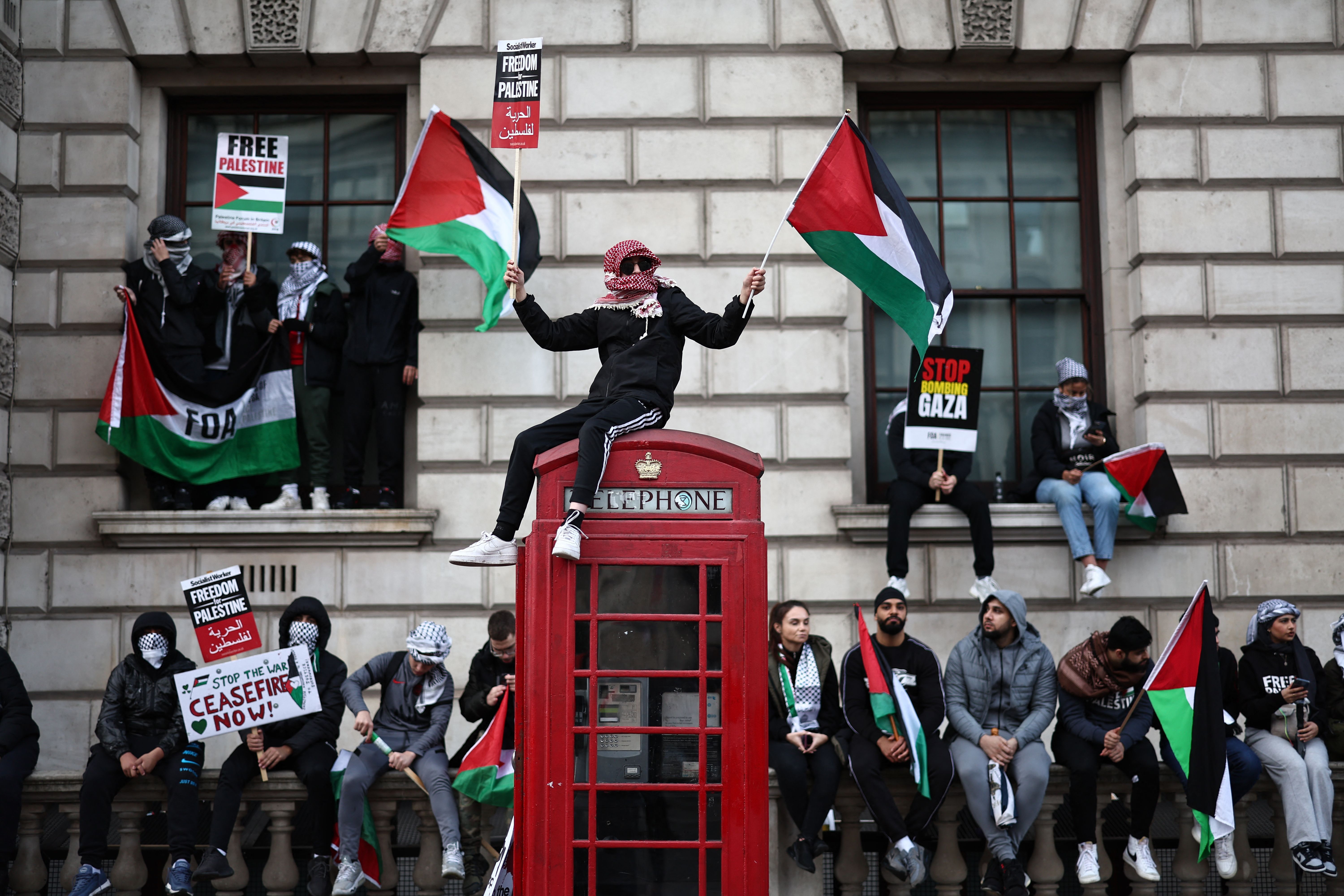 A protestor sits on a red telephone booth holding a Palestinian flag, surrounded by other protesters also holding flags and signs calling for a ceasefire. 