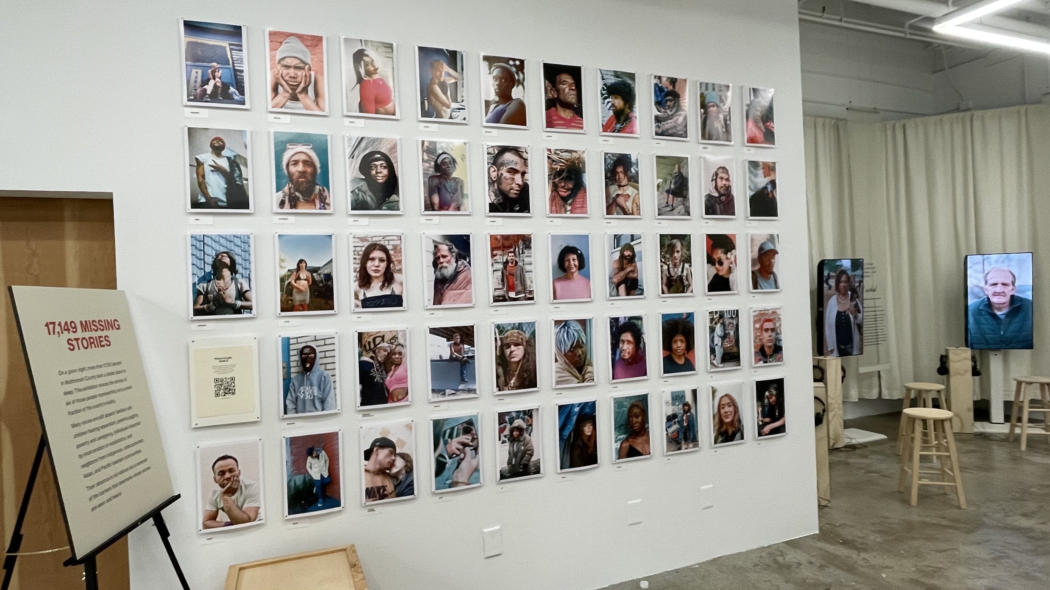 Art gallery wall displaying 48 colorful portraits of diverse individuals in six rows, next to a sign titled "17,149 MISSING STORIES" with explanatory text and QR code, and video screens with stools in the background.
