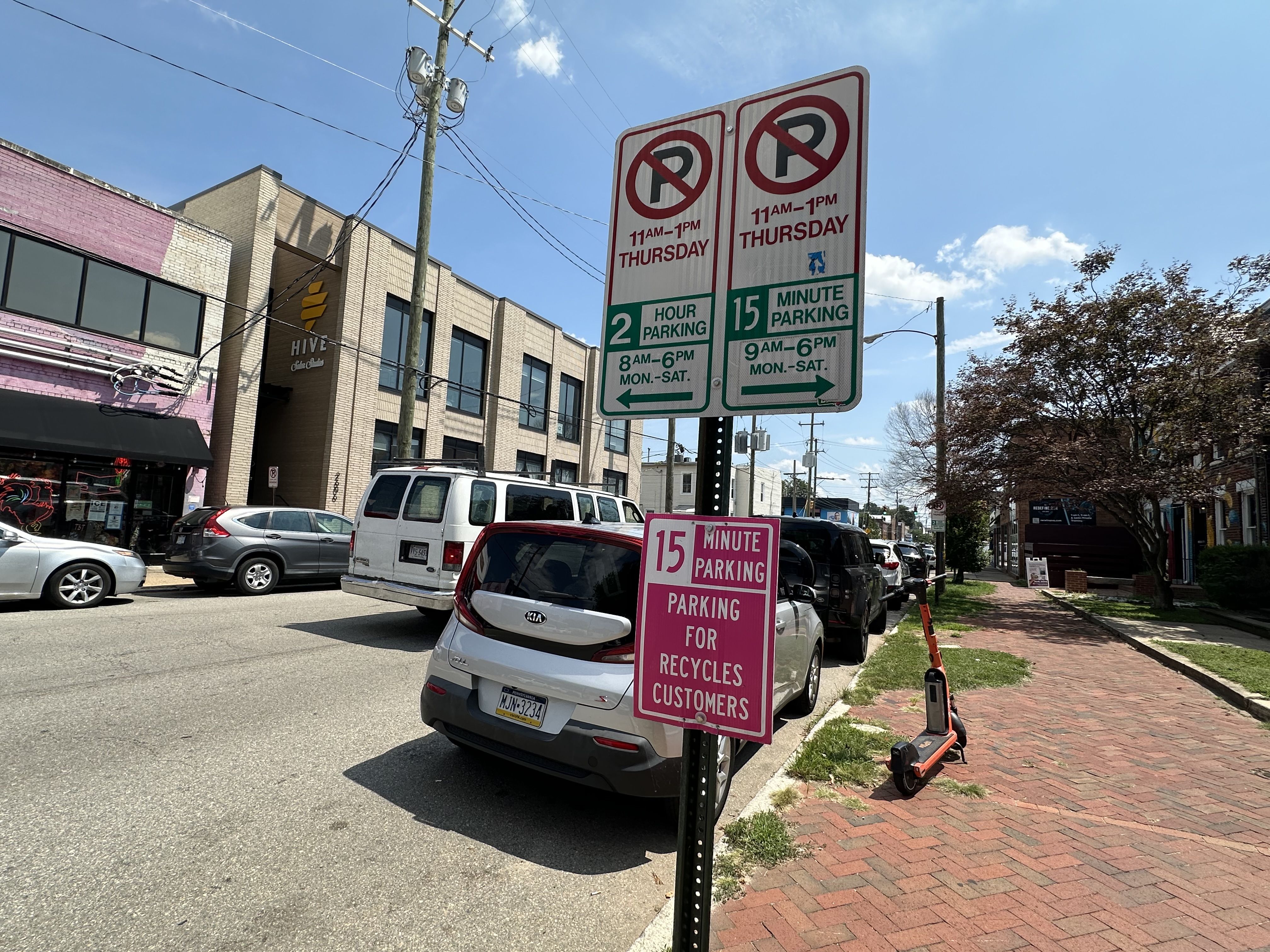 Urban street scene with a tall two-sided parking sign showing "No Parking" and hours; a foreground pink sign reads "15 Minute Parking for Customers"; brick sidewalk, parked cars, and blue sky.