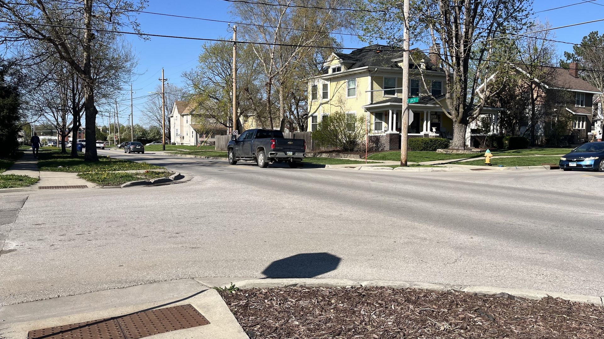 Suburban street corner with a pale yellow two-story house on the right, leafless trees, and overhead power lines. A black pickup crosses the intersection; a blue car is parked nearby.