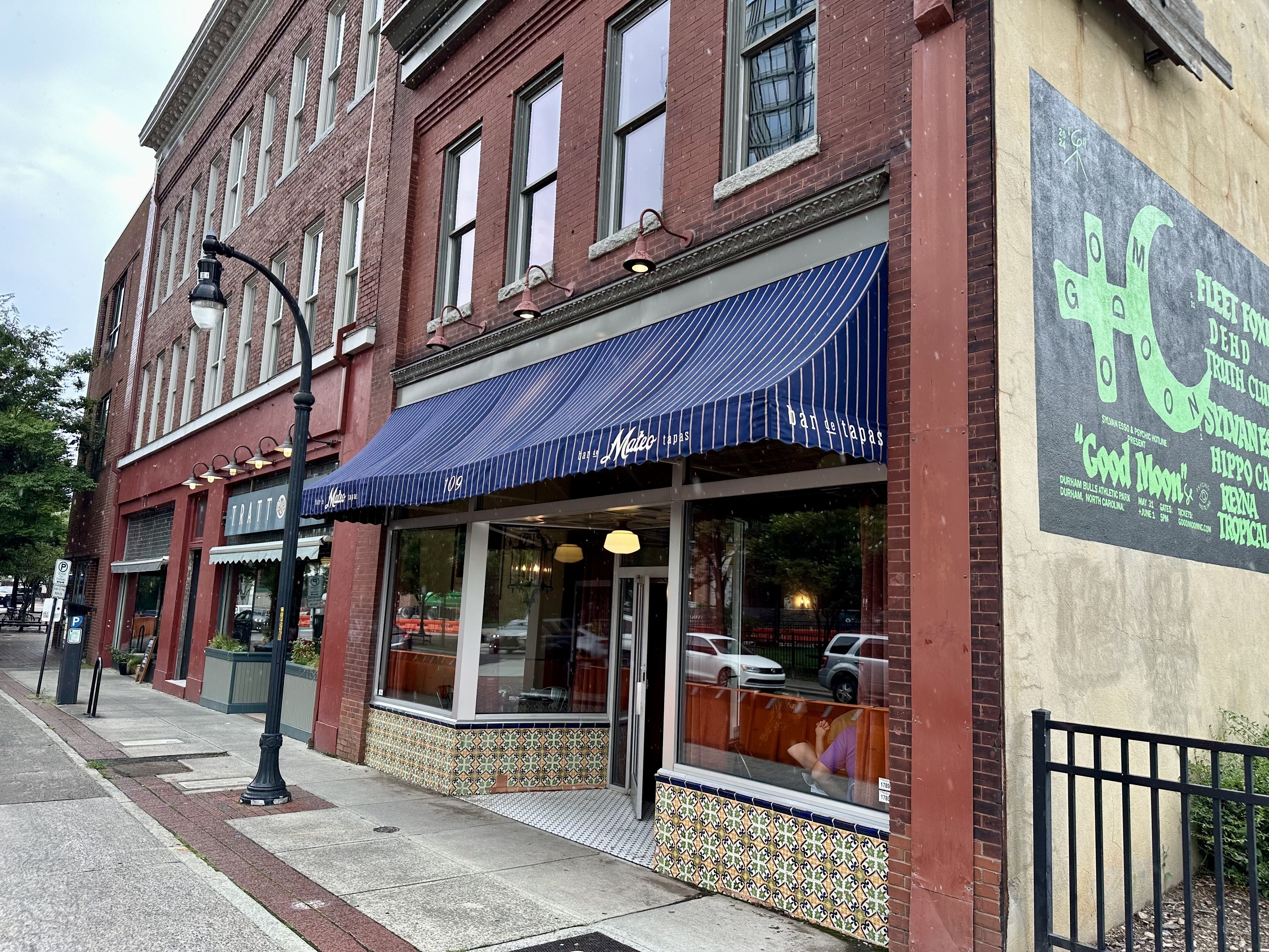 Urban street scene with a red brick building, tall windows, and a blue striped awning over a tapas shop. A curved streetlamp sits on the sidewalk; a green mural covers the side wall.