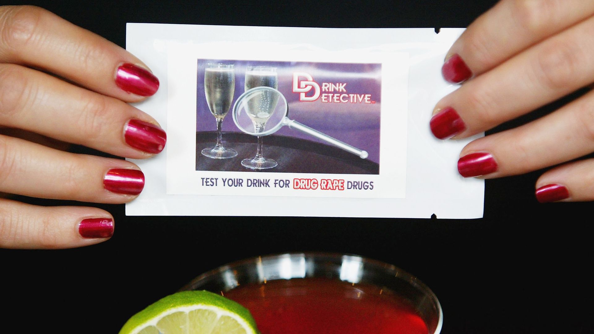 A pair of hands with fingernails painted red, holding a white rectangular package with the words "drink detective" above the words "test your drink for date rape drugs," over a glass with a red cocktail and a sliced lime on the rim.