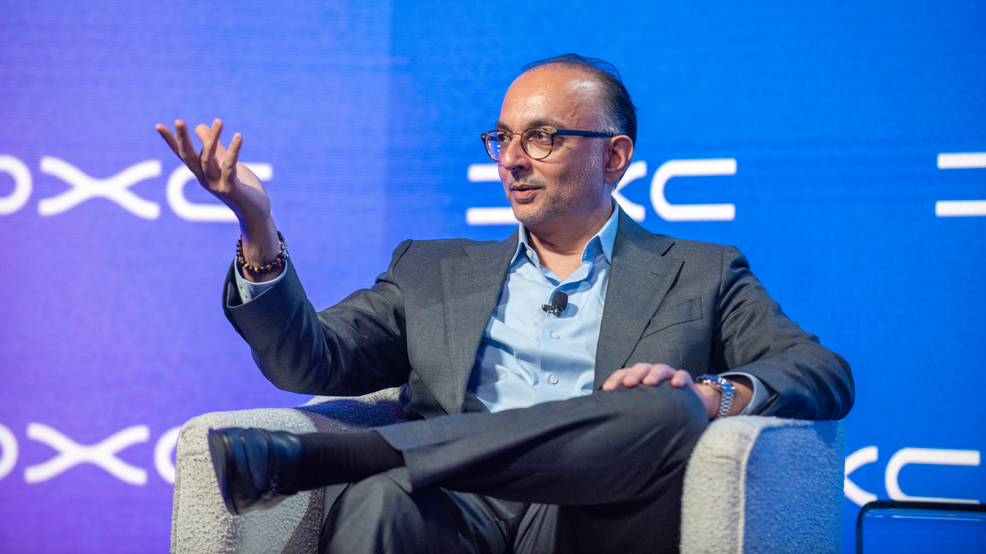 A man in a gray suit and light blue shirt sits on a stage, wearing glasses and a lapel mic, gesturing with his left hand against a blue backdrop.
