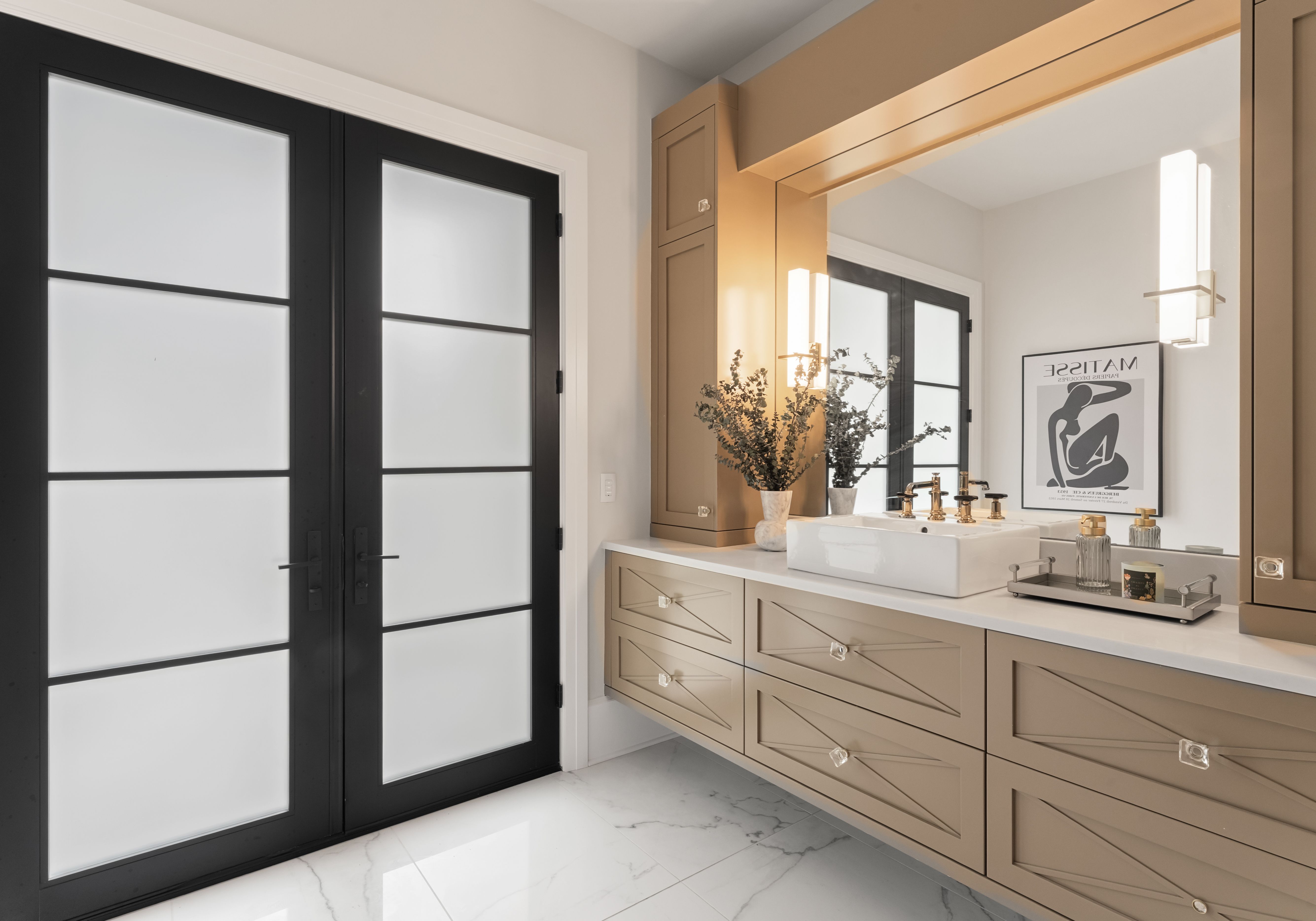 Modern bathroom with beige cabinets, large mirror, white rectangular sink with brass faucets, frosted black double doors, marble floor, and framed Matisse art. Vase with greenery on counter.