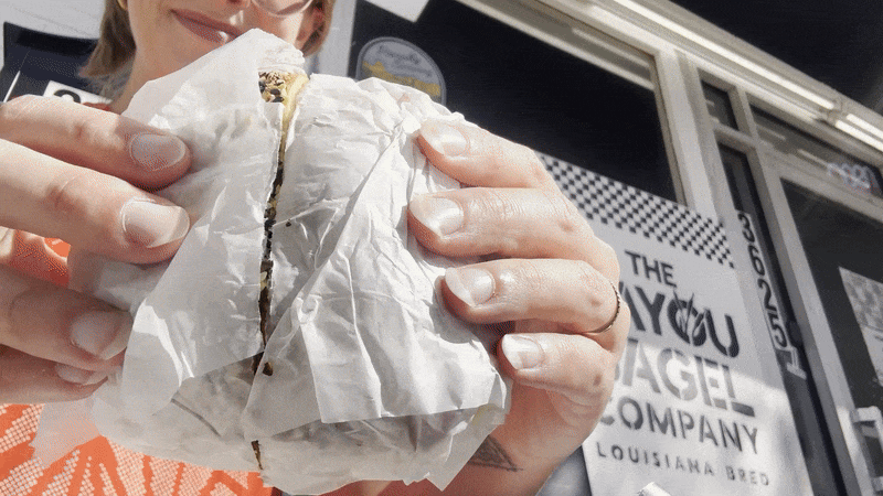 A partially obscured woman pulls apart a bagel sandwich with egg, bacon and cheese. A sign for The Bayou Bagel Company is on the storefront behind her.