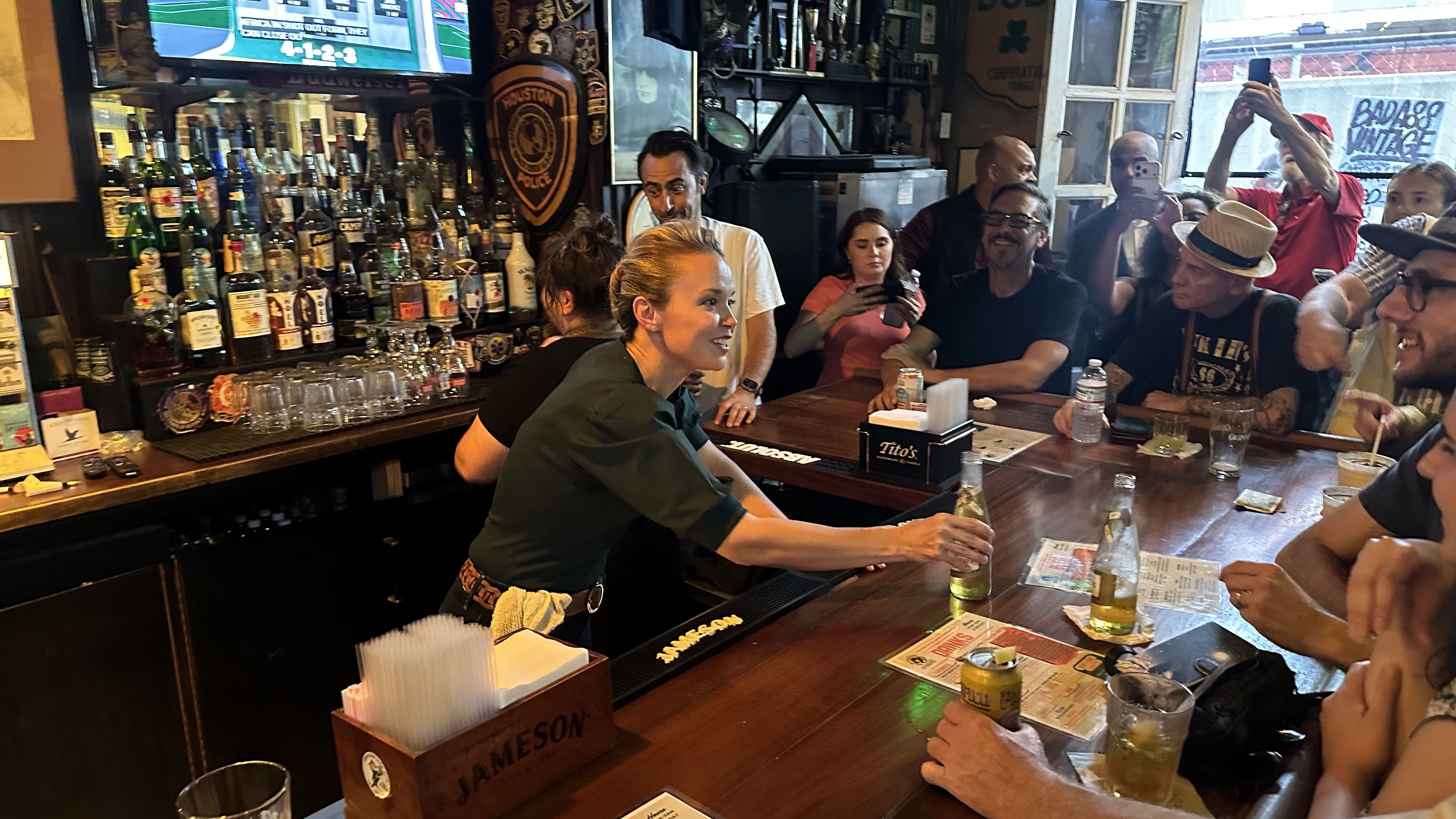 Lively bar interior with a smiling female bartender pouring a drink toward customers seated at a wooden counter; shelves of liquor line the back, while several people take photos.
