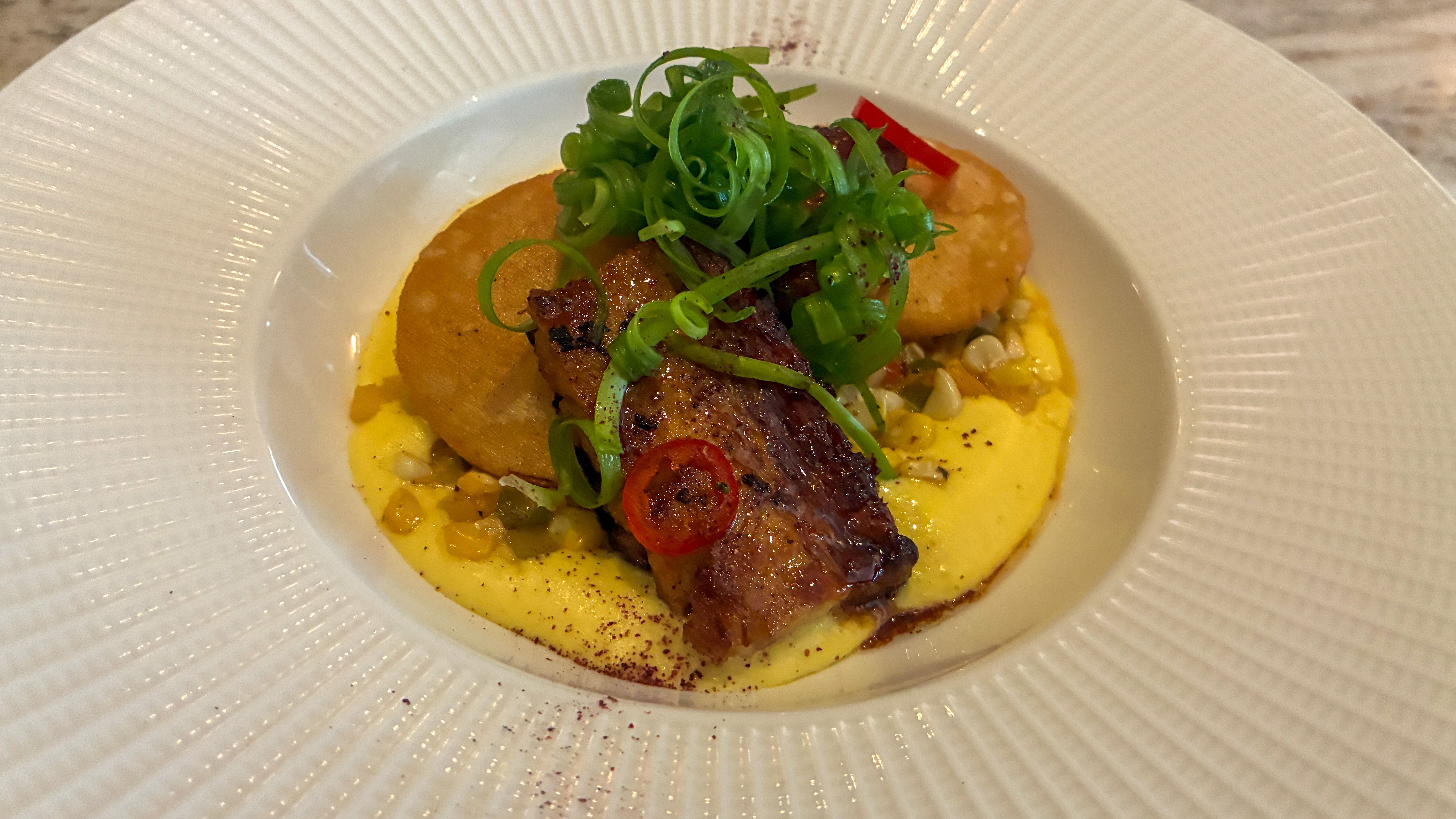 Seared meat atop a yellow creamy sauce with corn kernels and two golden polenta disks, garnished with green scallion curls and a red chili ring, served in a white textured bowl.